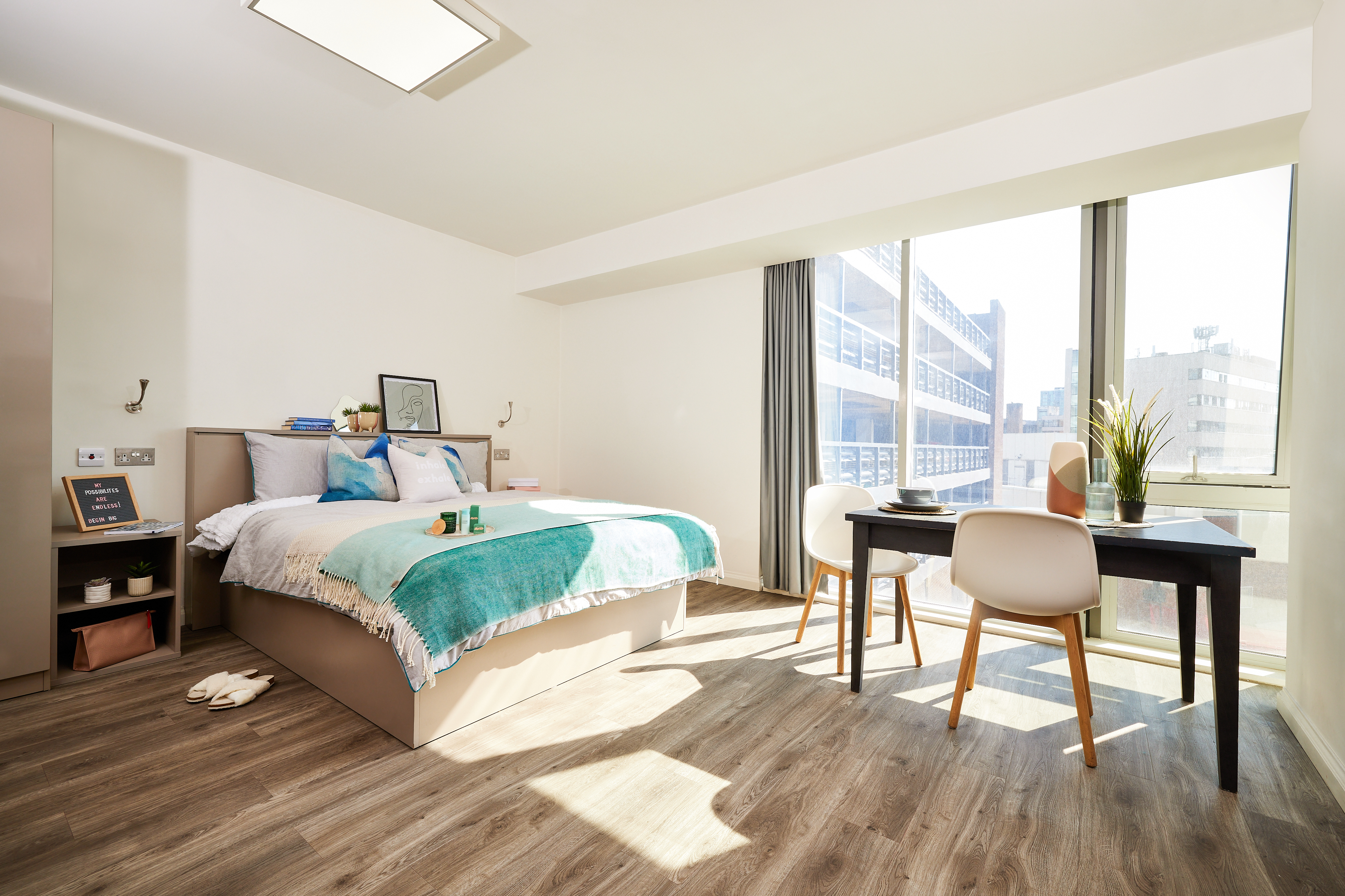 A bright, minimalist bedroom in Telephone House features a large window. The room includes a bed with teal and white bedding, a desk with two chairs, plants, and decorative items. Sunlight streams in through the window onto the wooden floor.
