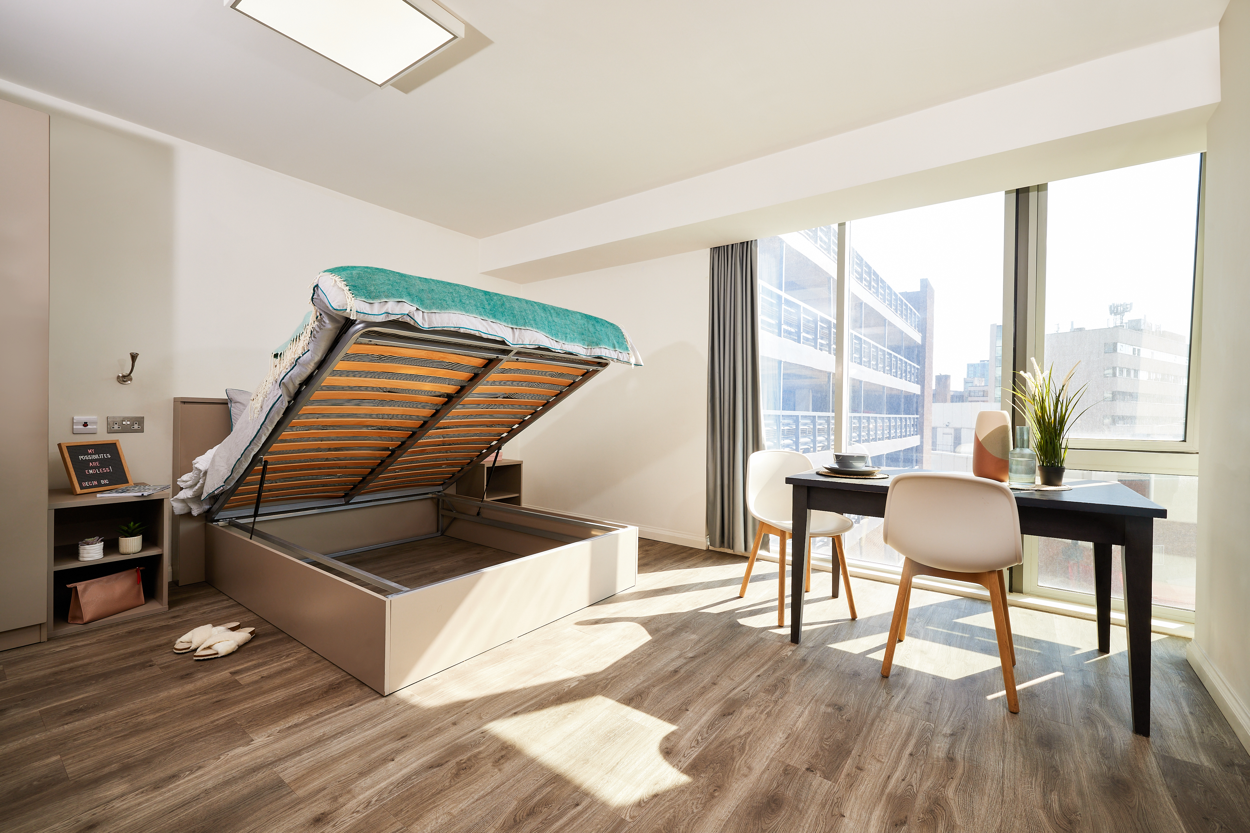 A modern studio apartment in Telephone House features a lifted storage bed revealing space underneath, two white chairs, and a dark wooden table by a large window with city views. The room boasts wood flooring, a potted plant, and bright natural light.
