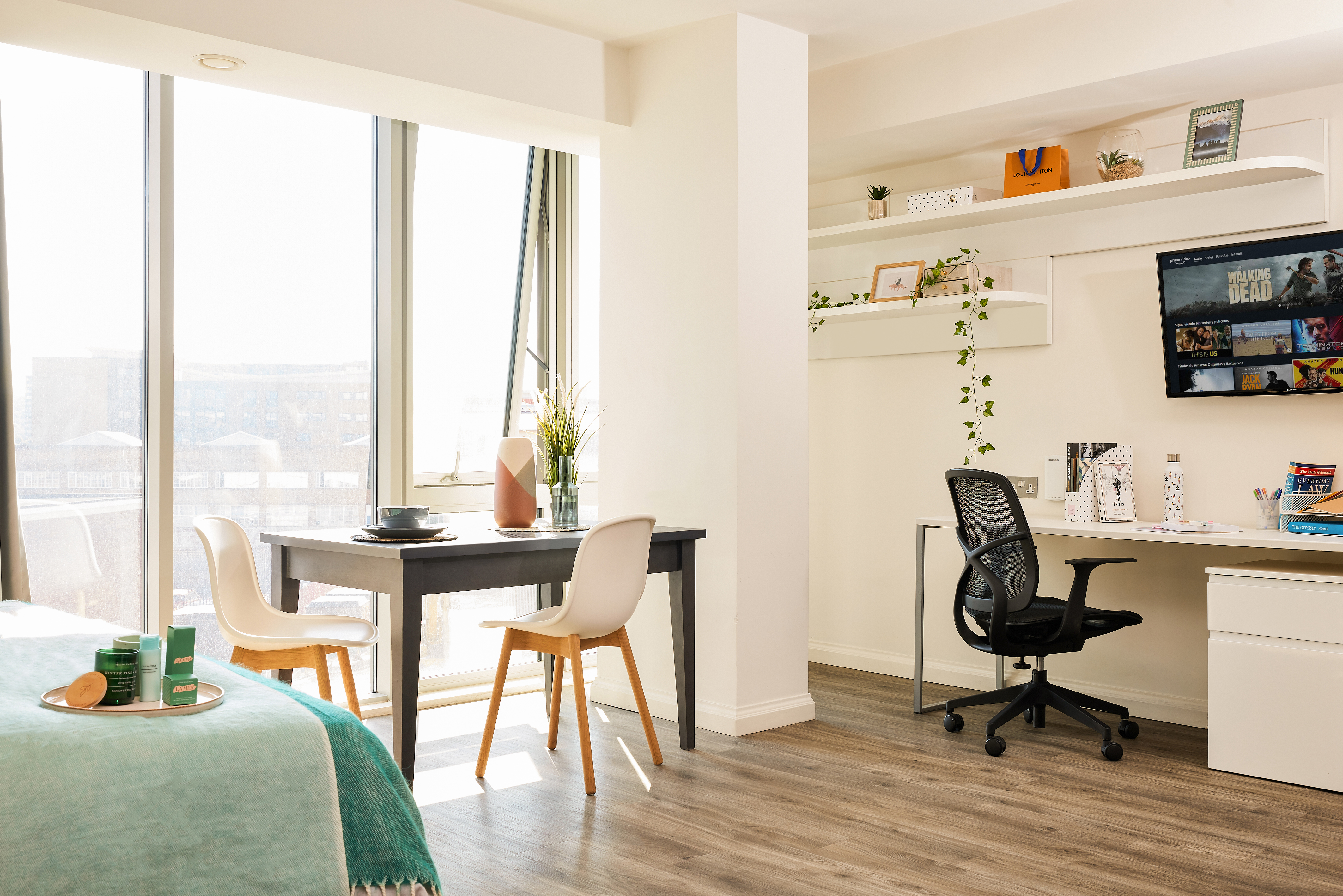 The bright apartment interior exudes warmth with its large window, dining table and chairs, and a thoughtfully organized workspace. A telephone sits neatly on the desk alongside a computer monitor and office chair. The cozy bed with a green blanket is partially visible as natural light illuminates the house-like ambiance.