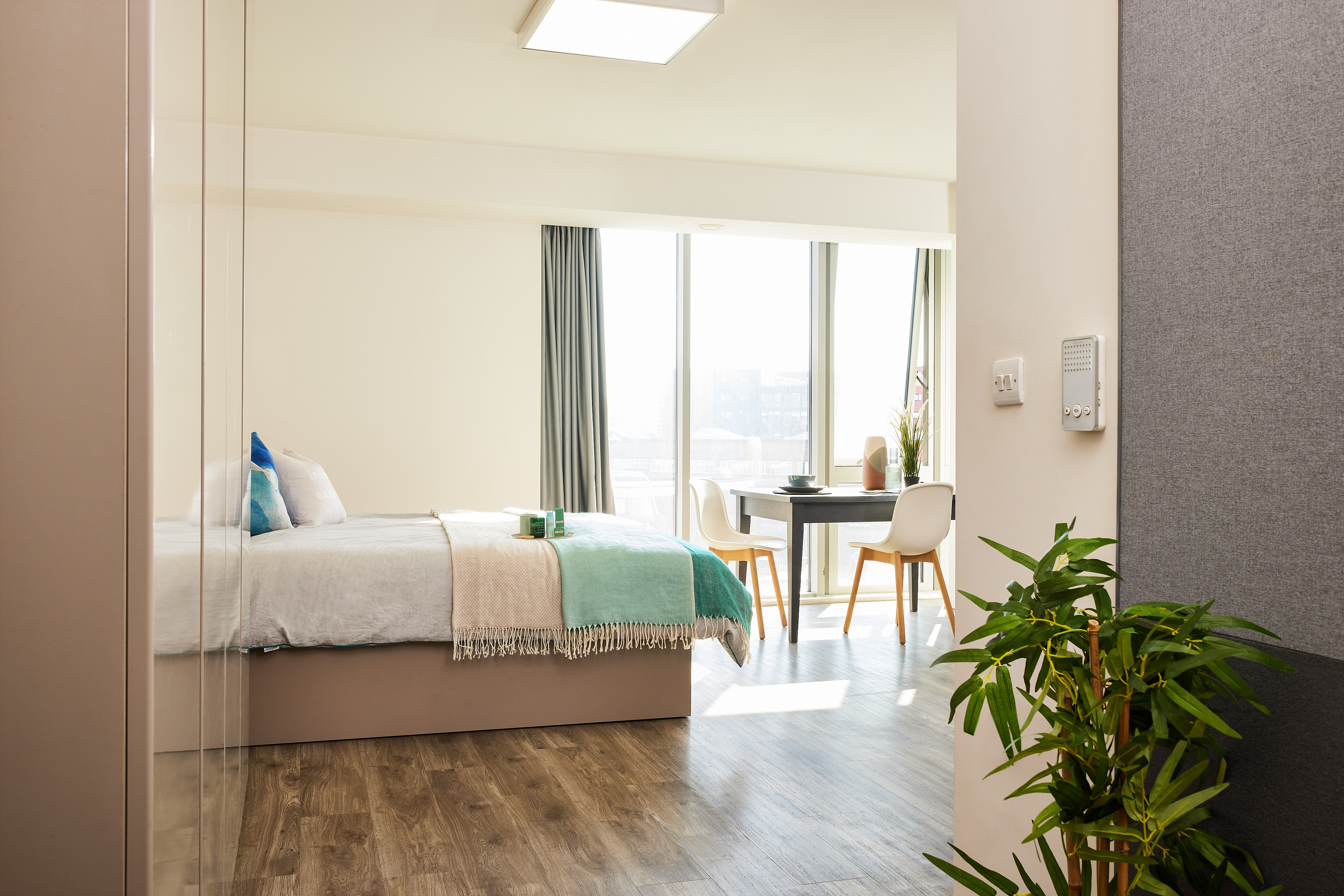 A bright bedroom with a neatly made bed featuring blue and beige bedding. Large windows let in natural light, and a small round dining table with two chairs is in the corner. A green plant sits beside an antique telephone, adding charm to the cozy space.
