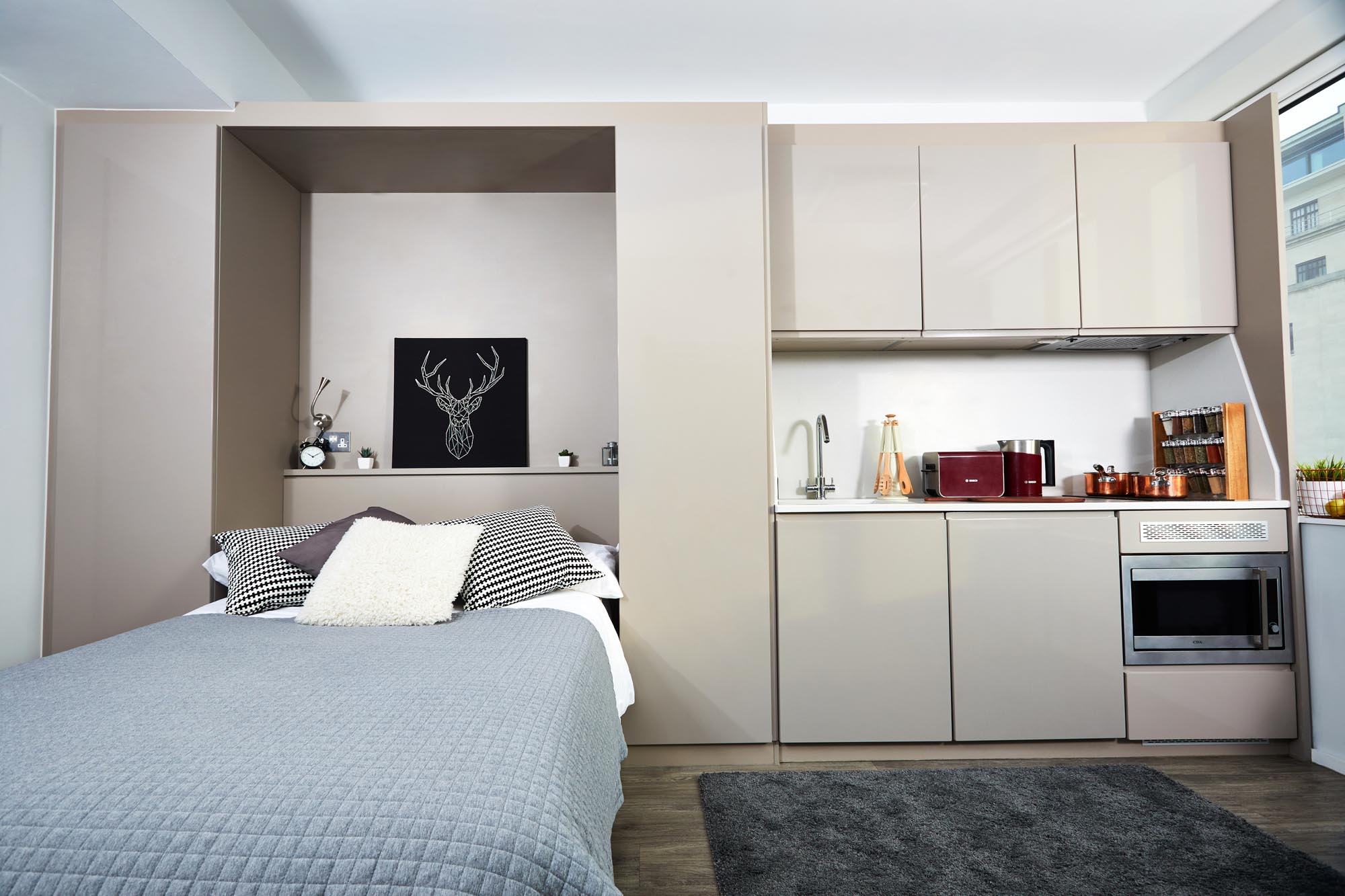 A compact studio apartment featuring a bed with gray bedding on the left and a small kitchen area on the right. The kitchen includes cabinets, a sink, microwave, and appliances. A deer artwork is above the bed, and a window offers city views.