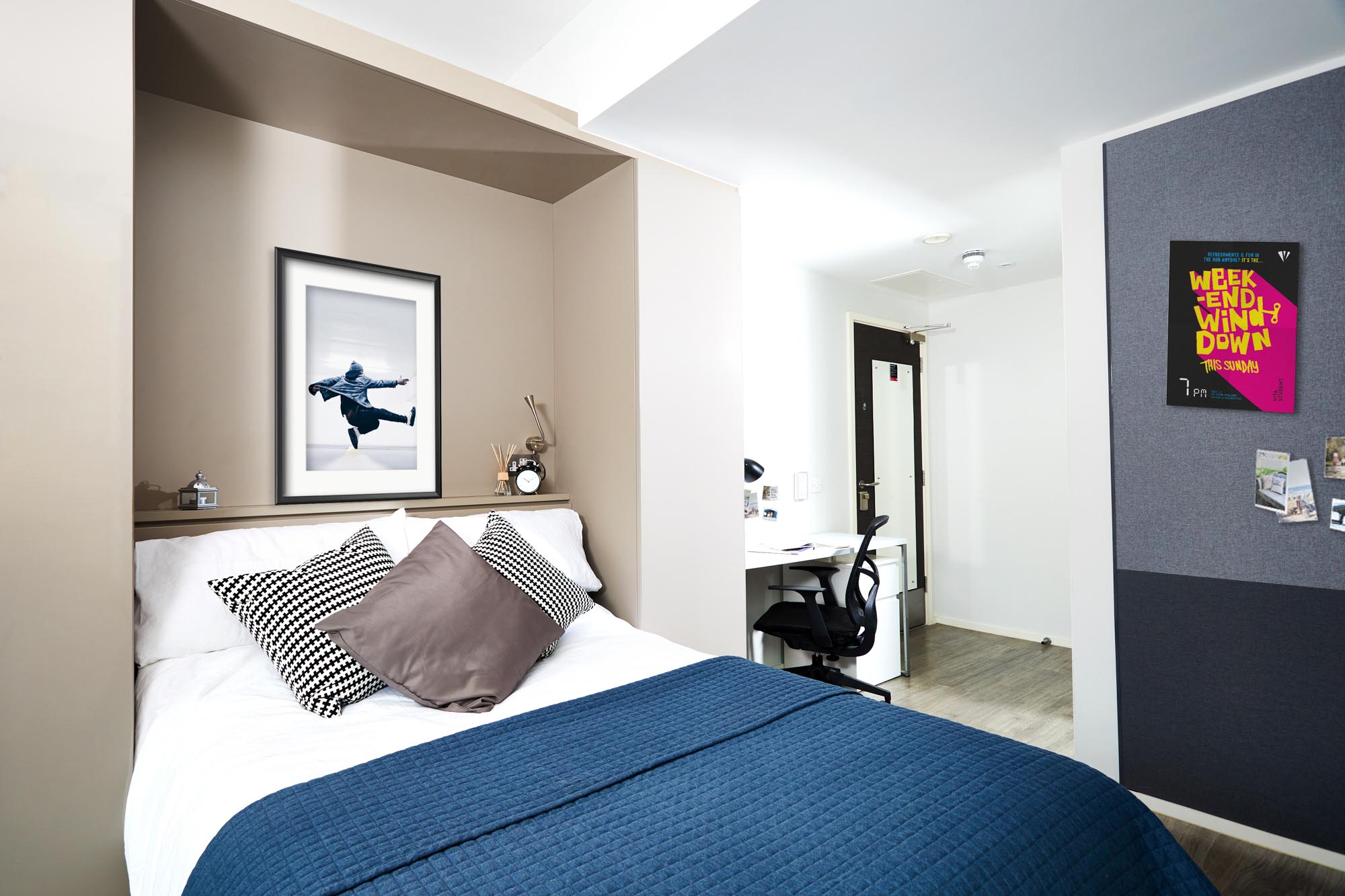 A modern bedroom with a bed featuring a blue quilt and several pillows, against a beige accent wall with a framed photograph. A desk and chair are visible in the background, and a colorful poster is on the wall.