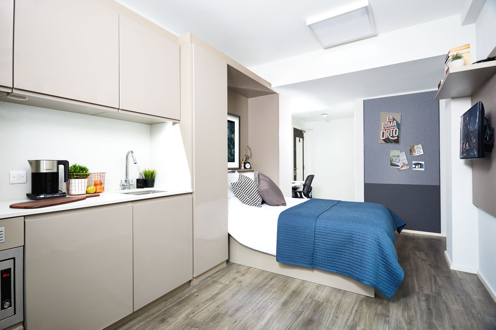 Modern studio apartment with a small kitchen featuring light cabinetry, sink, microwave, and coffee maker. Adjacent is a bed with a blue blanket and pillows. A desk with a chair is in the corner. Walls are decorated with artwork and shelves.