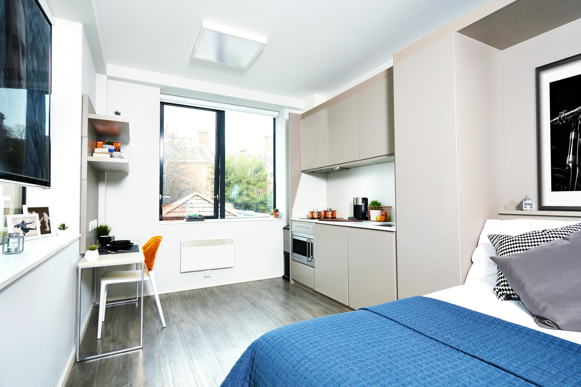 A modern studio apartment with a bed, blue bedding, a compact kitchen area, a desk with a chair, and a window with a view of trees. The room has light-colored walls, wood flooring, and is decorated with framed art.