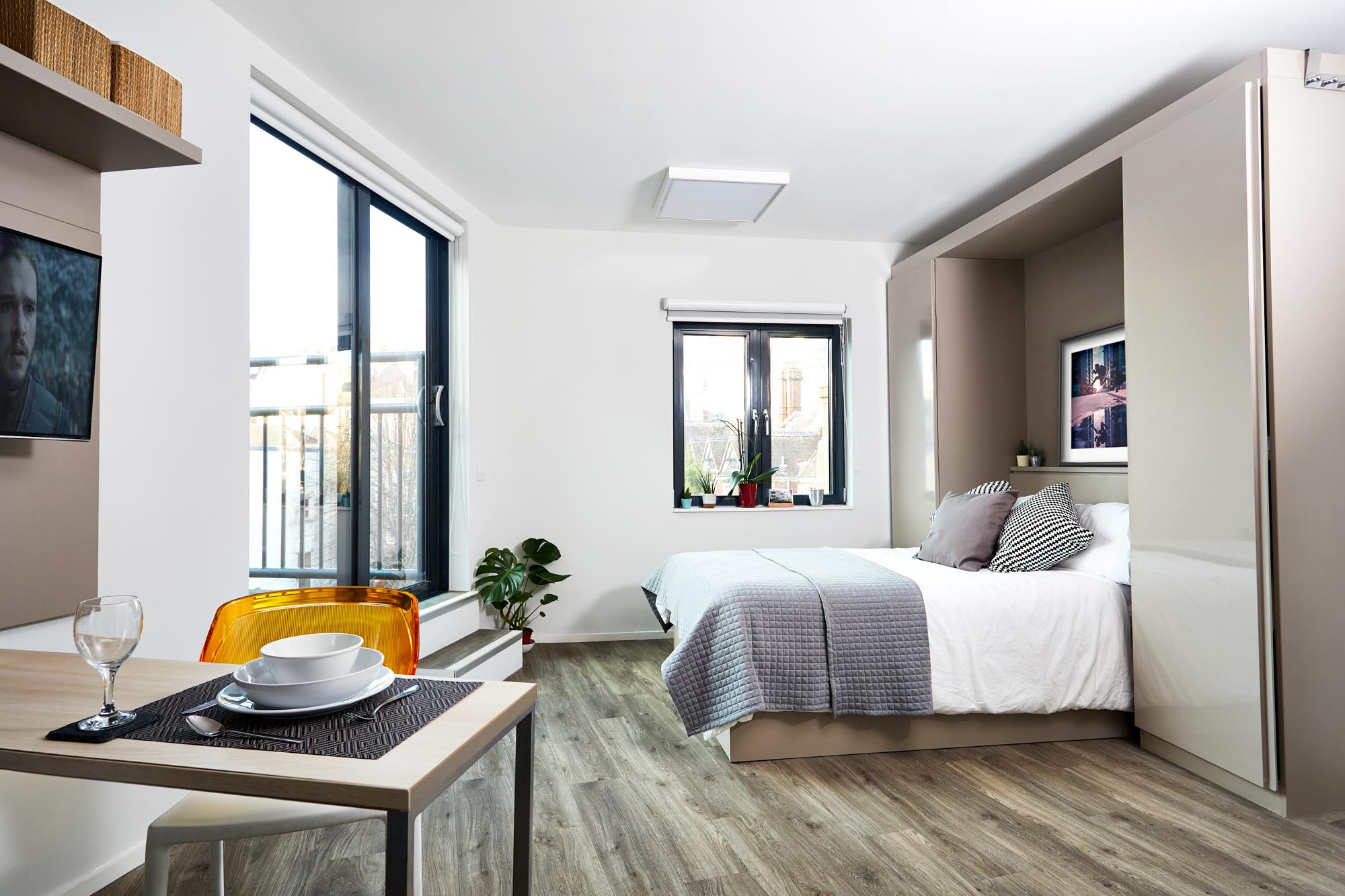 Modern studio apartment with a foldable bed, gray bedding, a small dining table set for two, and a clear chair. Large windows provide natural light. The walls are white, and the flooring is wood, with a potted plant near the window.