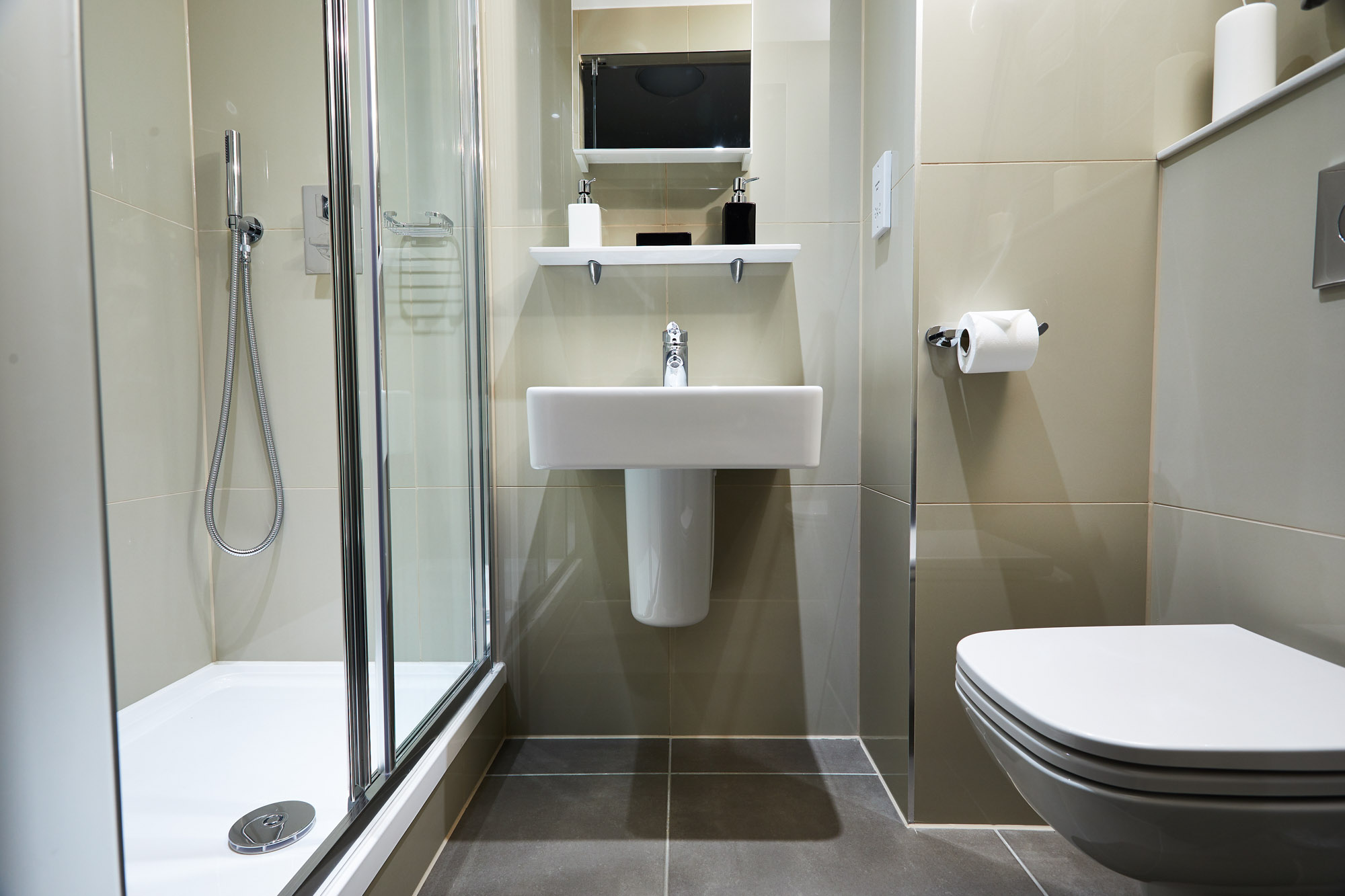 Modern bathroom with a glass shower enclosure on the left, a white pedestal sink in the center with a mirror above, and a wall-mounted toilet on the right. Neutral beige and gray tiles cover the walls and floor.