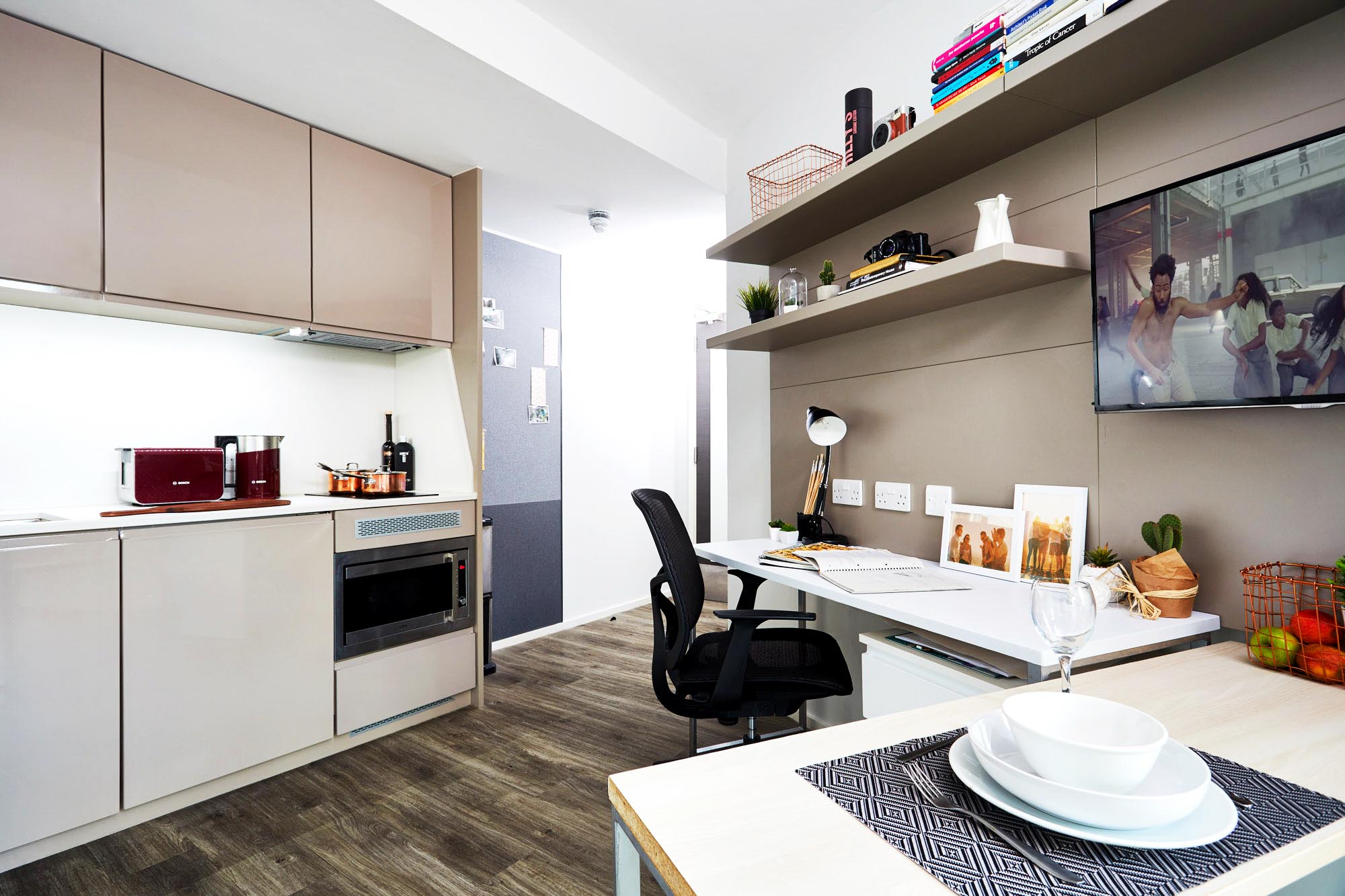 Modern studio apartment with a compact kitchen featuring beige cabinets, a microwave, and a kettle. A sleek desk with a lamp and office chair is adjacent, below wall-mounted shelves. A TV is on the wall, and a small dining table is in the foreground.