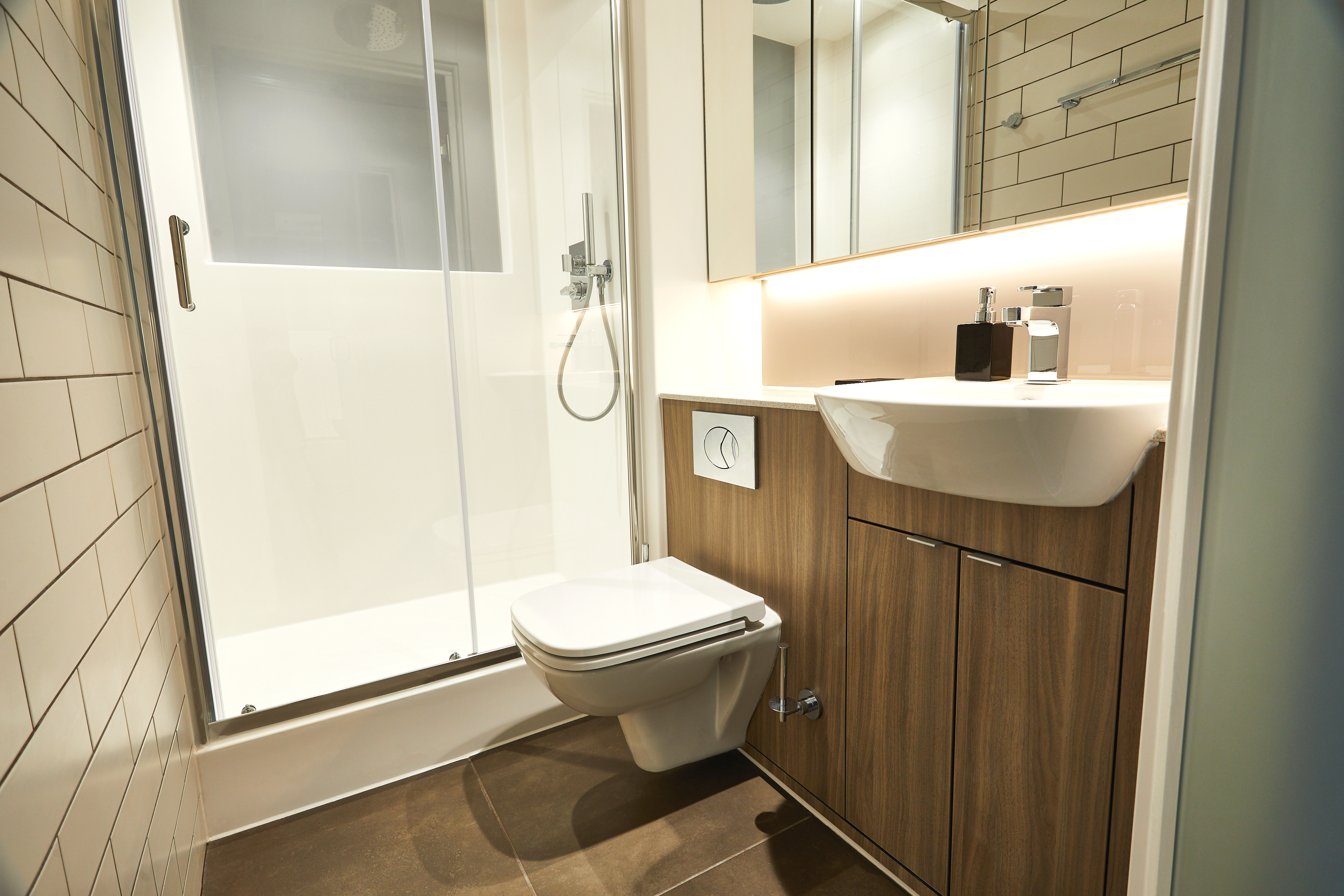 Modern bathroom with a glass-enclosed shower, wall-mounted toilet, and wood-paneled vanity. The sink is integrated into the vanity, topped with a mirror and illuminated by soft lighting. Beige tiles line one wall and the floor.