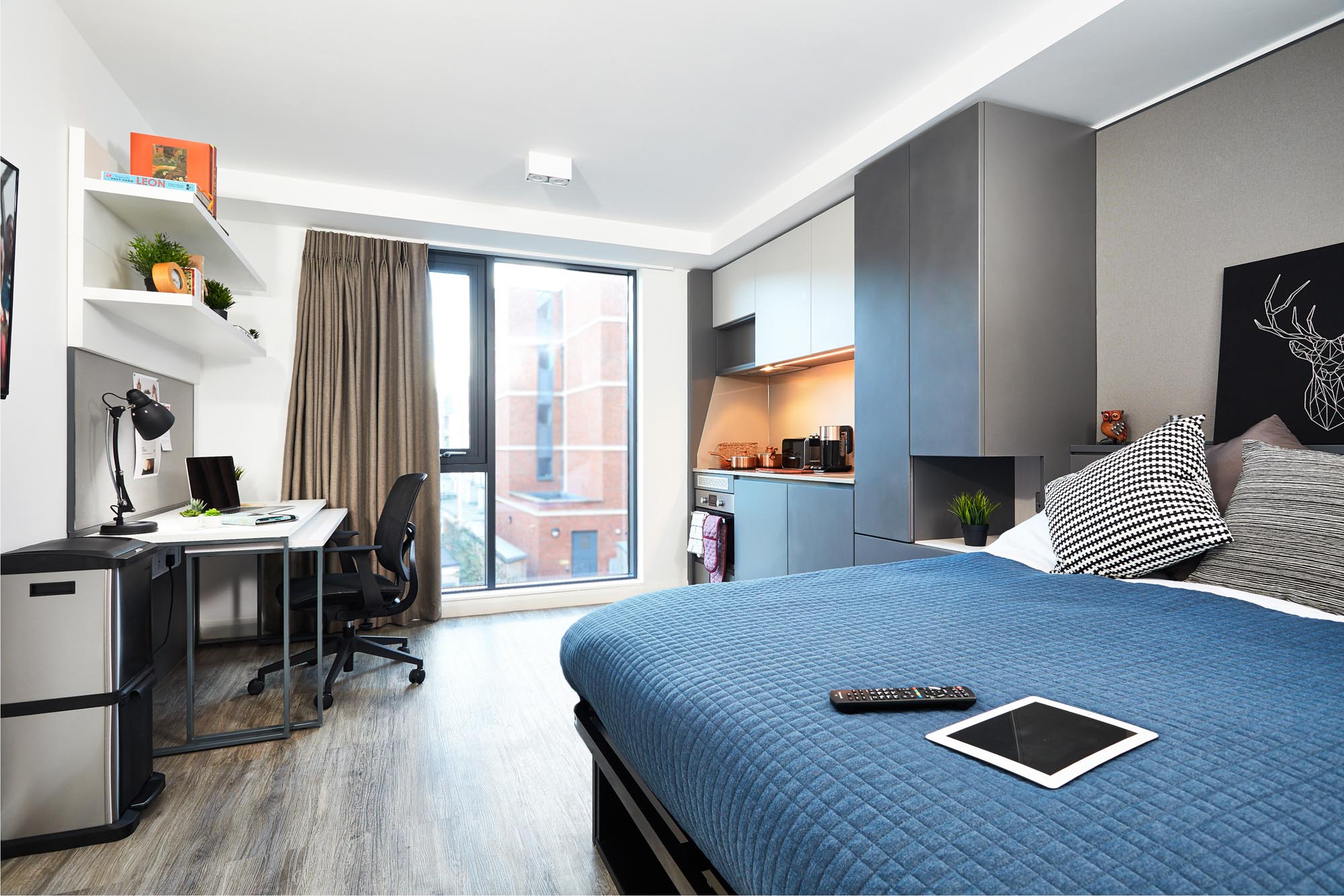 Modern studio apartment with a bed covered in a blue quilt, a tablet and remote on the bed, and a kitchen area with a microwave. A desk with a chair, lamp, and shelves is near the window, which overlooks a brick building.