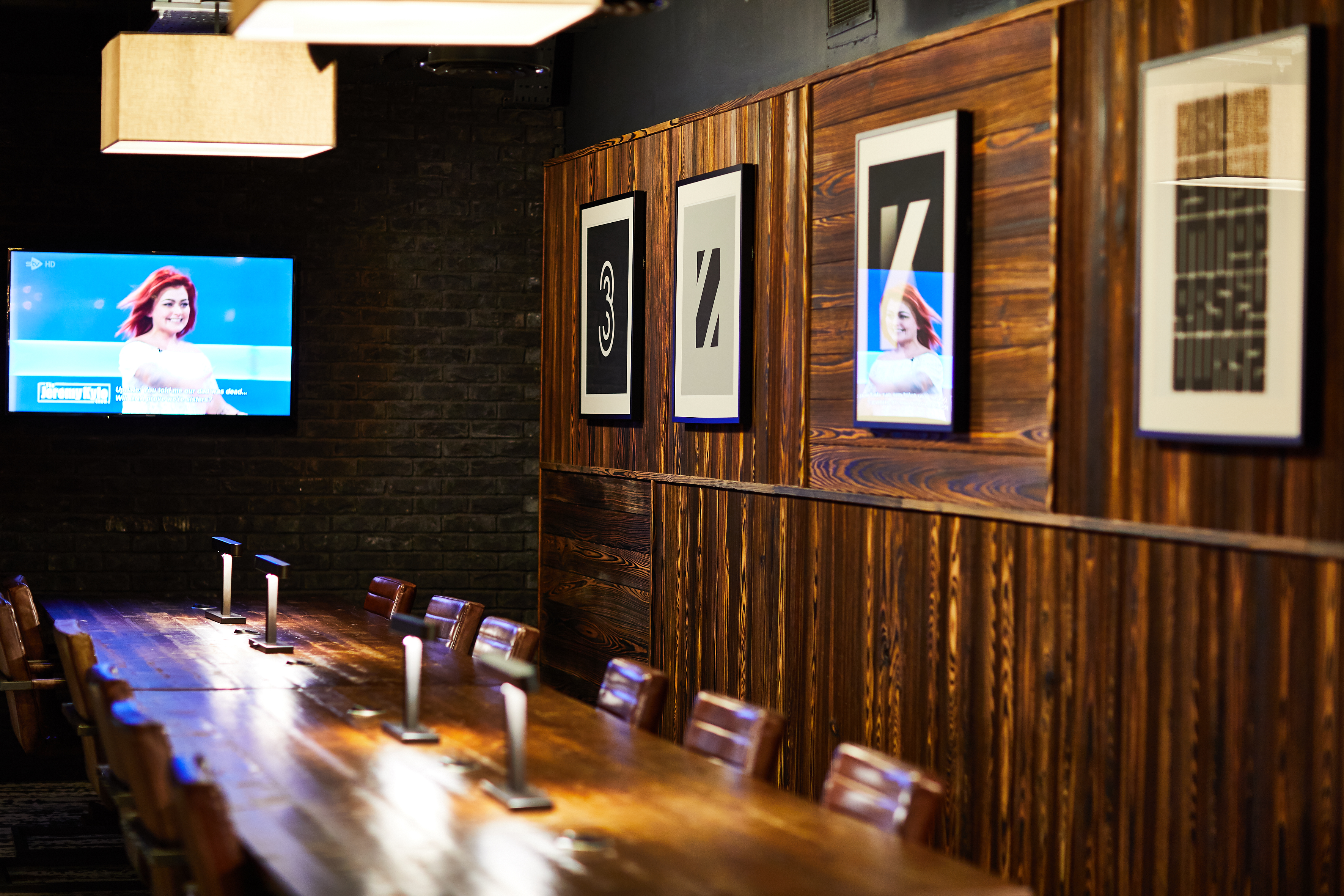 A dimly lit dining room in Fountainbridge features a long wooden table and chairs. Wooden walls showcase several framed abstract artworks. A TV on the left displays an image of a person with red hair, while hanging lamps cast warm light over the cozy setting.