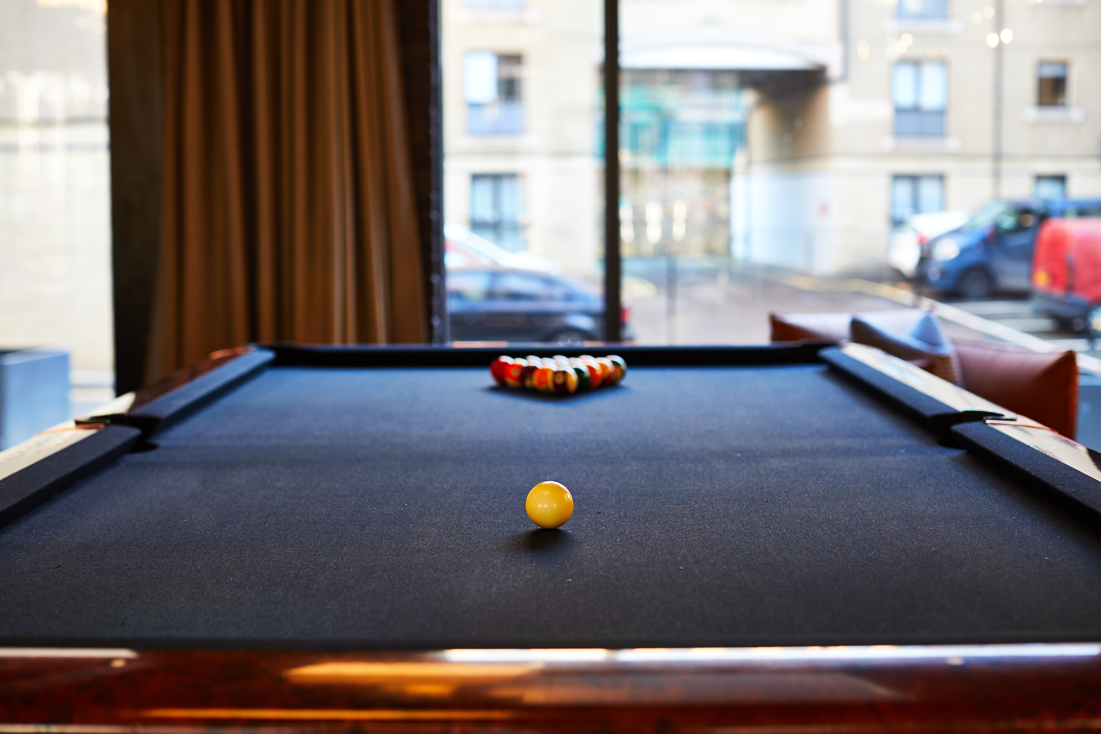 A pool table with a yellow cue ball in the foreground and a triangle of colored balls at the far end sits in a room at Fountainbridge. Large windows offer an exterior view of a building and parked cars, adding to the vibrant atmosphere.