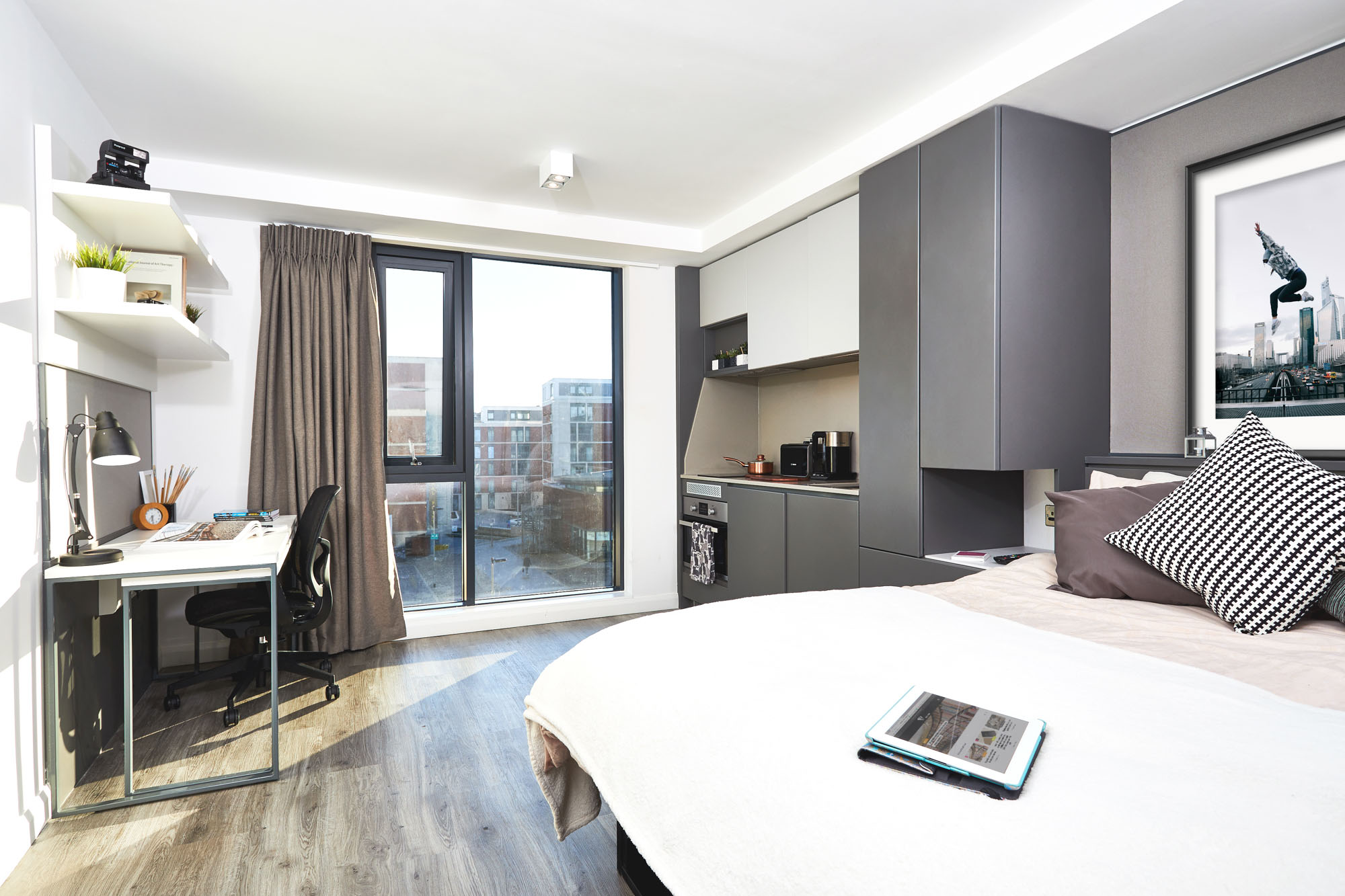 In the heart of Fountainbridge, this modern, bright bedroom boasts a large window with stunning city views. The room features a minimalist style with wooden floors, a cozy bed with pillows, a desk complete with stationery and laptop, and a sleek wall-mounted shelf for added elegance.