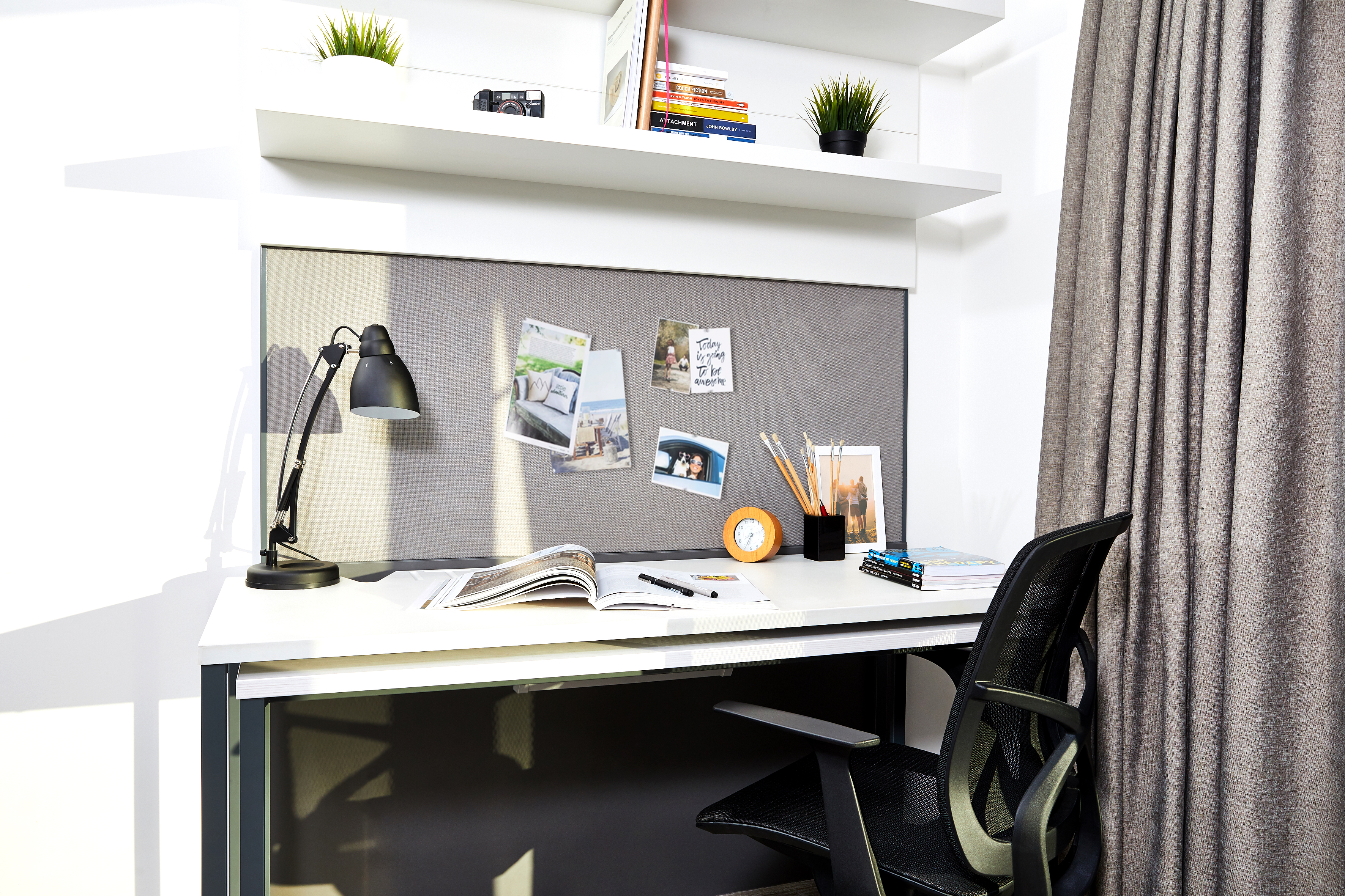 A tidy desk setup in Fountainbridge features a white surface, black lamp, and open book. A pinboard displays photos and pins. Shelves above hold books and plants. An orange clock, pencil holder, and books are also on the desk. A black chair is partially visible.