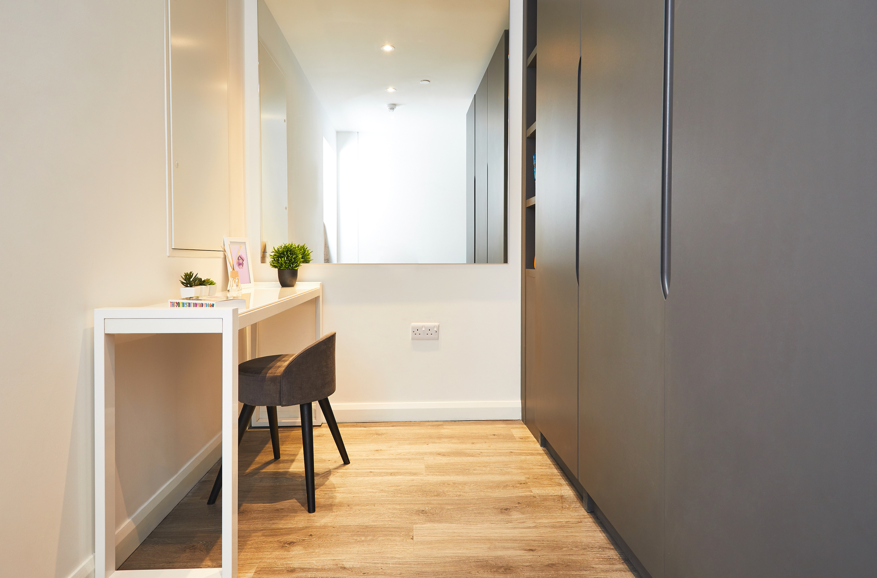 A minimalist room in Fountainbridge with a wooden floor, featuring a white vanity desk and a black chair on the left. A large mirror reflects the space. On the right, dark cabinets stand tall. Small plants and a picture frame adorn the desk, adding a touch of elegance.