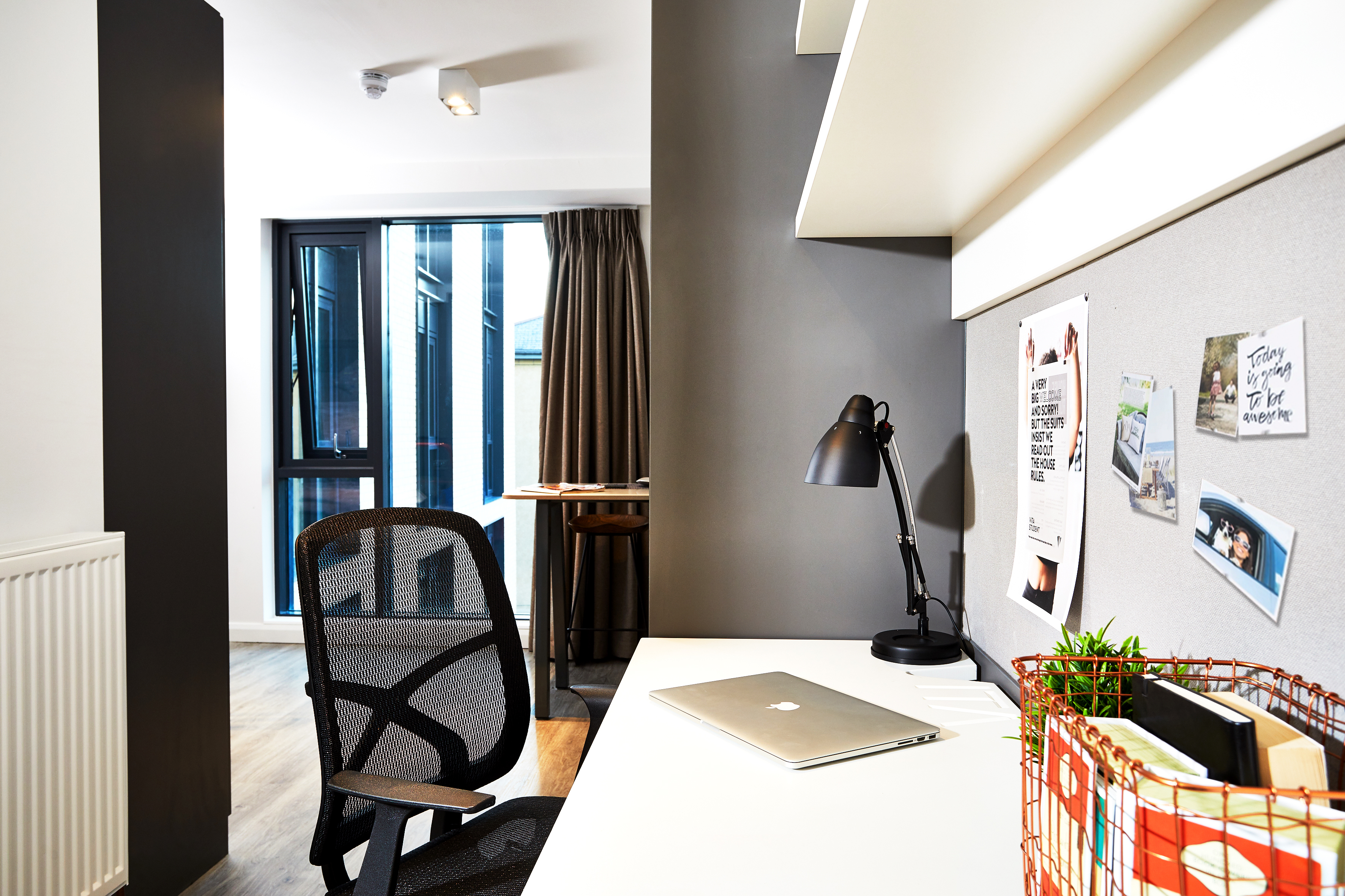 A modern study area in Fountainbridge features a desk with a laptop, desk lamp, and notes pinned on a gray board. Beside the desk is a mesh office chair. Large windows with curtains allow natural light to brighten the space.