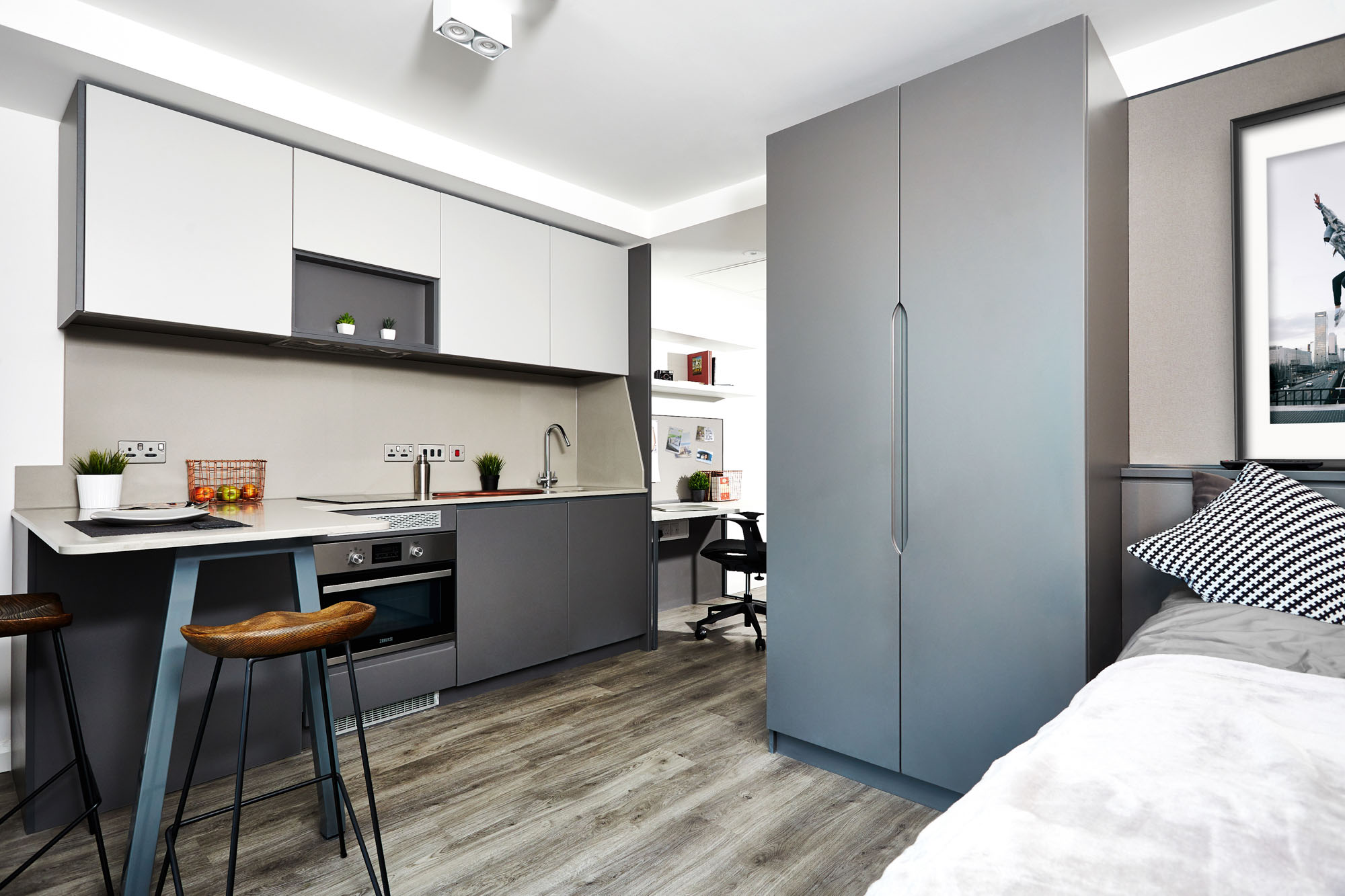 Modern studio apartment in Fountainbridge with a sleek kitchen featuring white and gray cabinets, an oven, and bar stools. A compact workspace with a desk and chair is visible. Light wood flooring, a large wardrobe, and a bed with monochrome bedding complete the space.