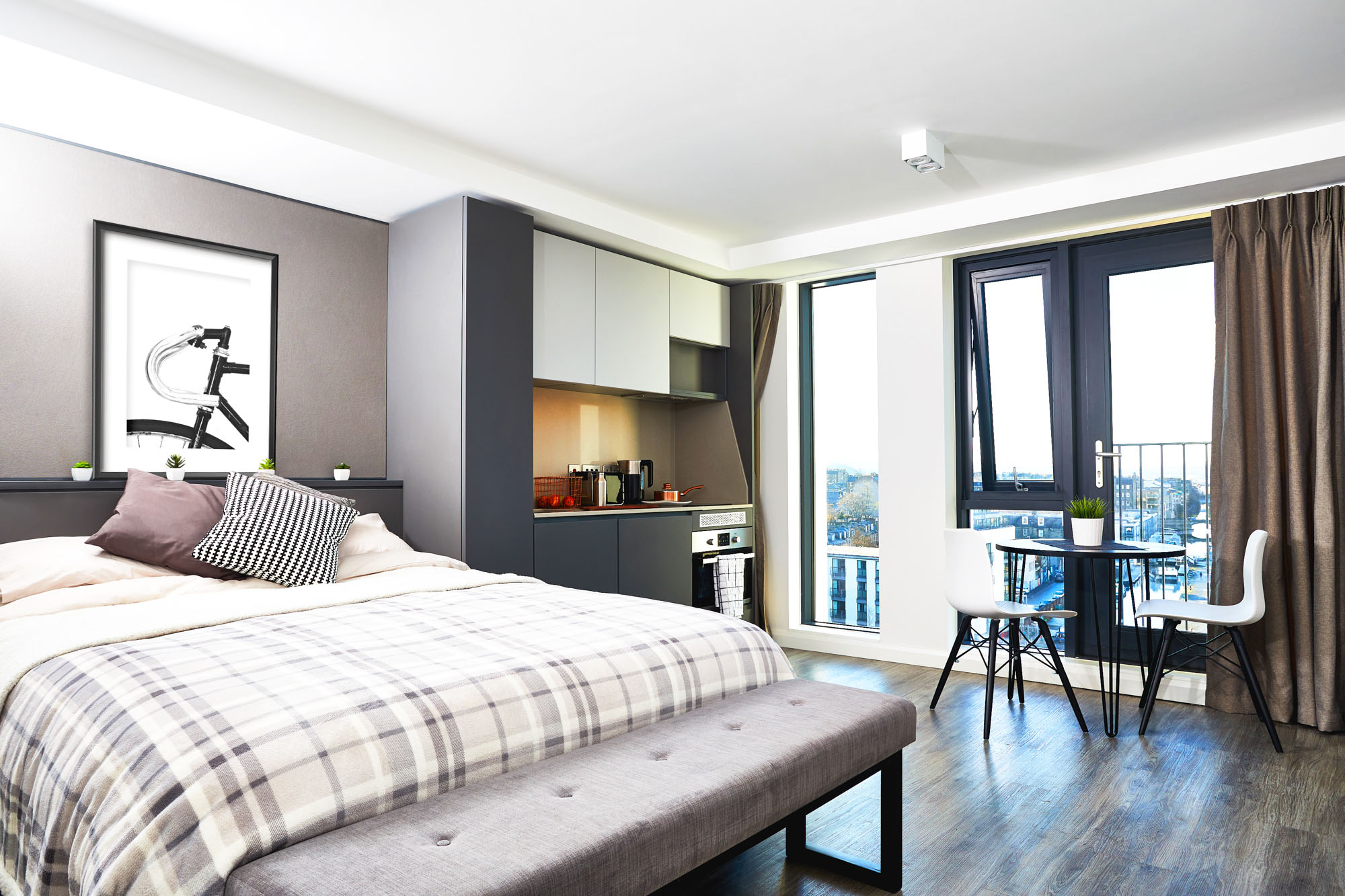 Modern studio apartment in Fountainbridge featuring a neatly made bed with a plaid blanket and cushions. A small kitchen area includes cabinets, stove, and kettle. A round table with two chairs sits near large windows with curtains, letting in an abundance of natural light.