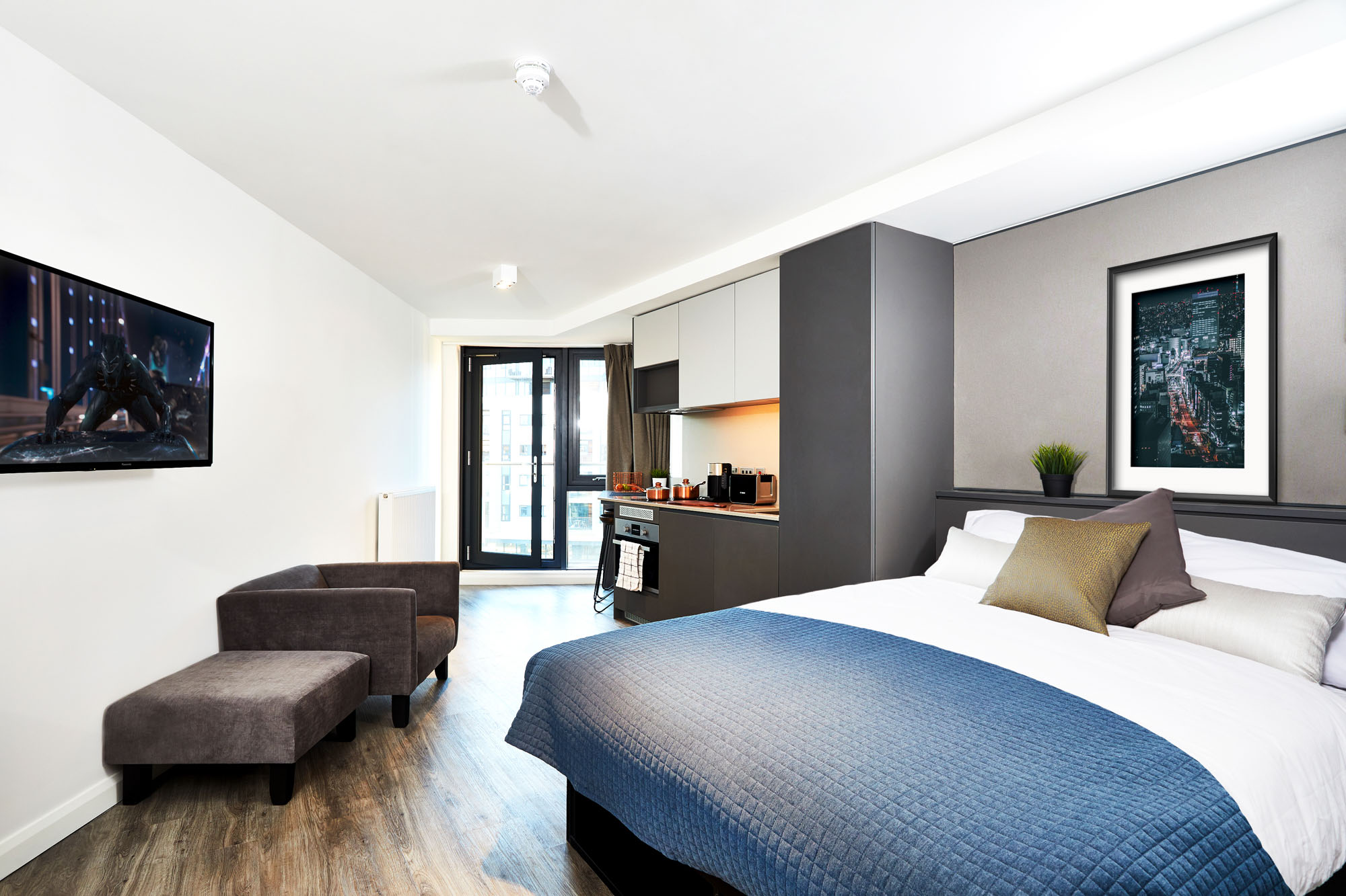 Located in the lively area of Fountainbridge, this modern studio apartment boasts a bed with a blue quilt, gray headboard, and assorted pillows. The kitchenette includes cabinets and a built-in oven. Enjoy entertainment on the wall-mounted TV by the large window with two brown chairs nearby.