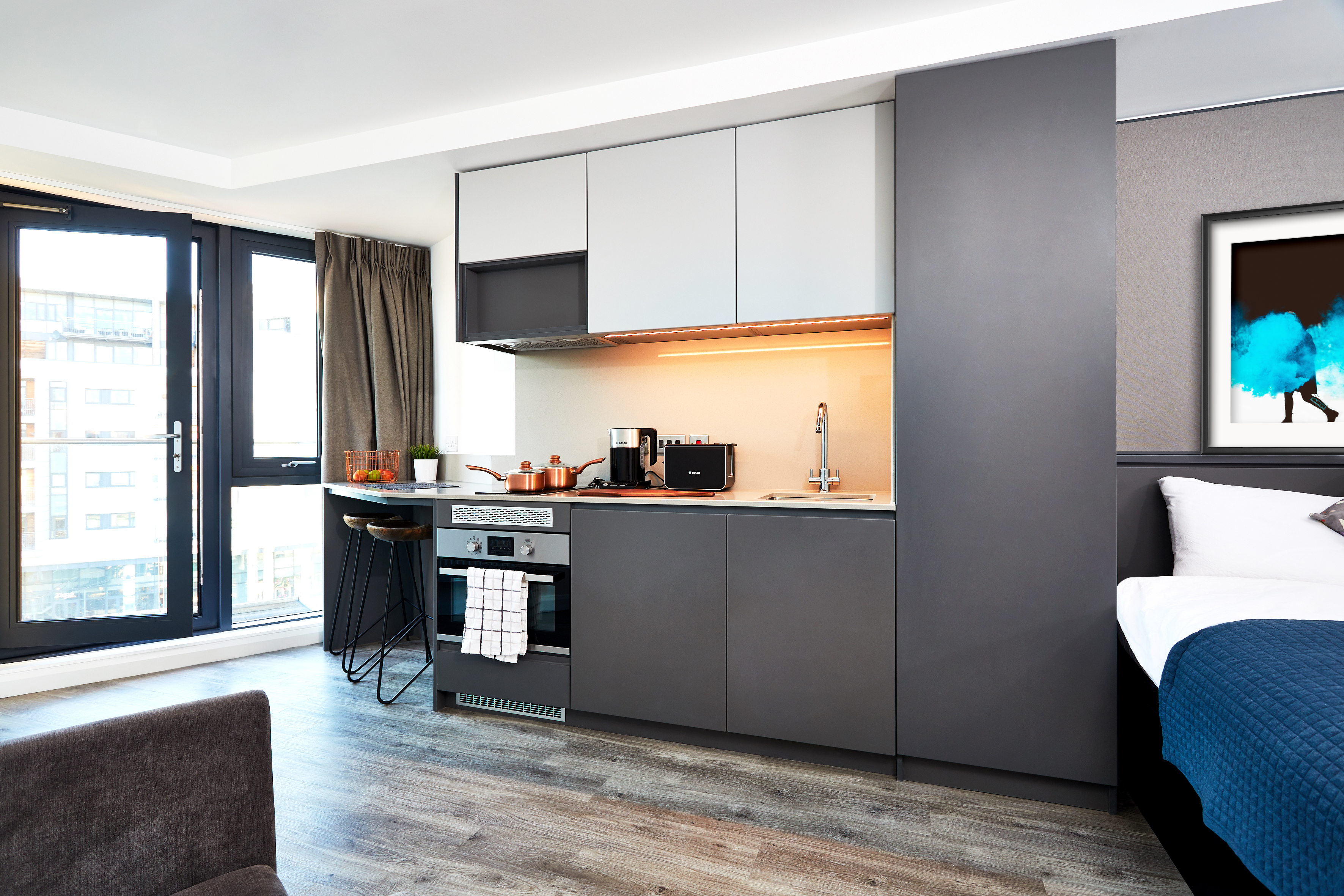 Modern studio apartment in Fountainbridge with a sleek kitchen featuring dark cabinets, a built-in oven, and small appliances. A table with stools sits by large windows. The room includes a bed with blue bedding, while a vibrant abstract painting adorns the wall.