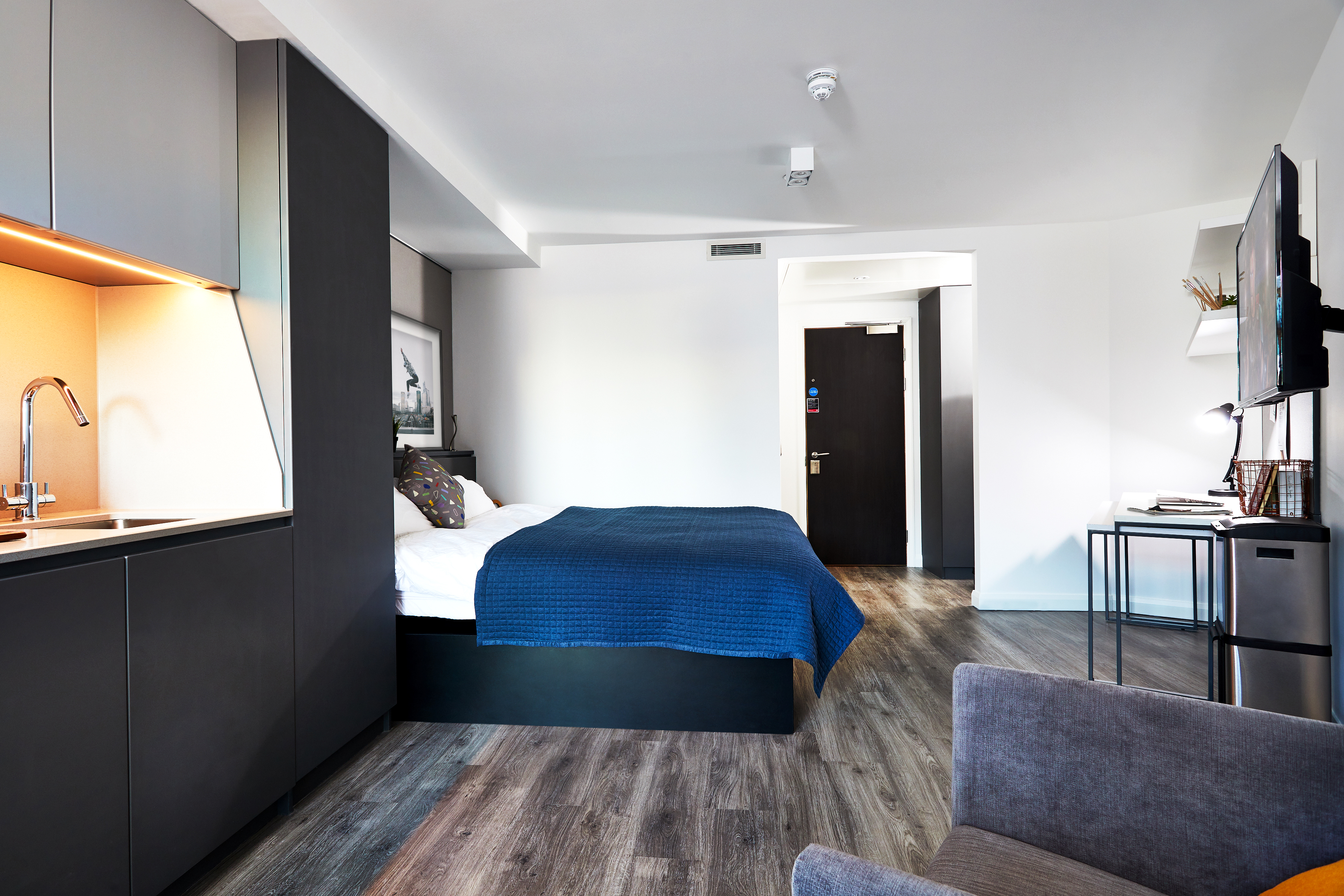 Nestled in Fountainbridge, this modern studio apartment features a small kitchenette, a bed with a blue throw, and a wall-mounted TV. The room boasts wood flooring, a gray armchair, and a desk adorned with decorative items by the entryway.
