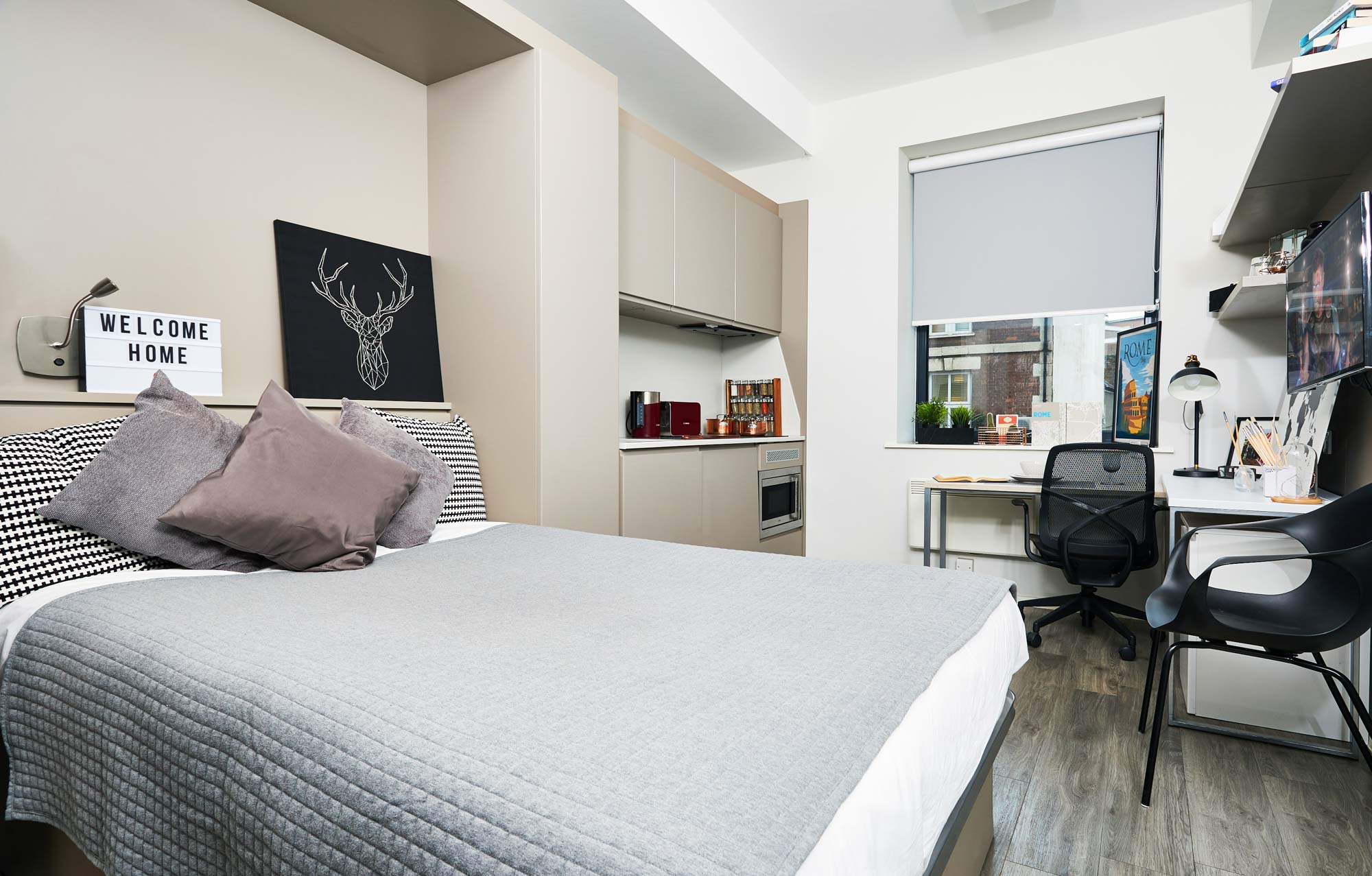 Modern studio apartment in Portland with a neatly made bed and decorative pillows. A "Welcome Home" sign greets you, complemented by a small kitchenette with appliances, a desk with a chair and lamp, and a window featuring a roller blind. The wooden flooring enhances the minimalistic decor.