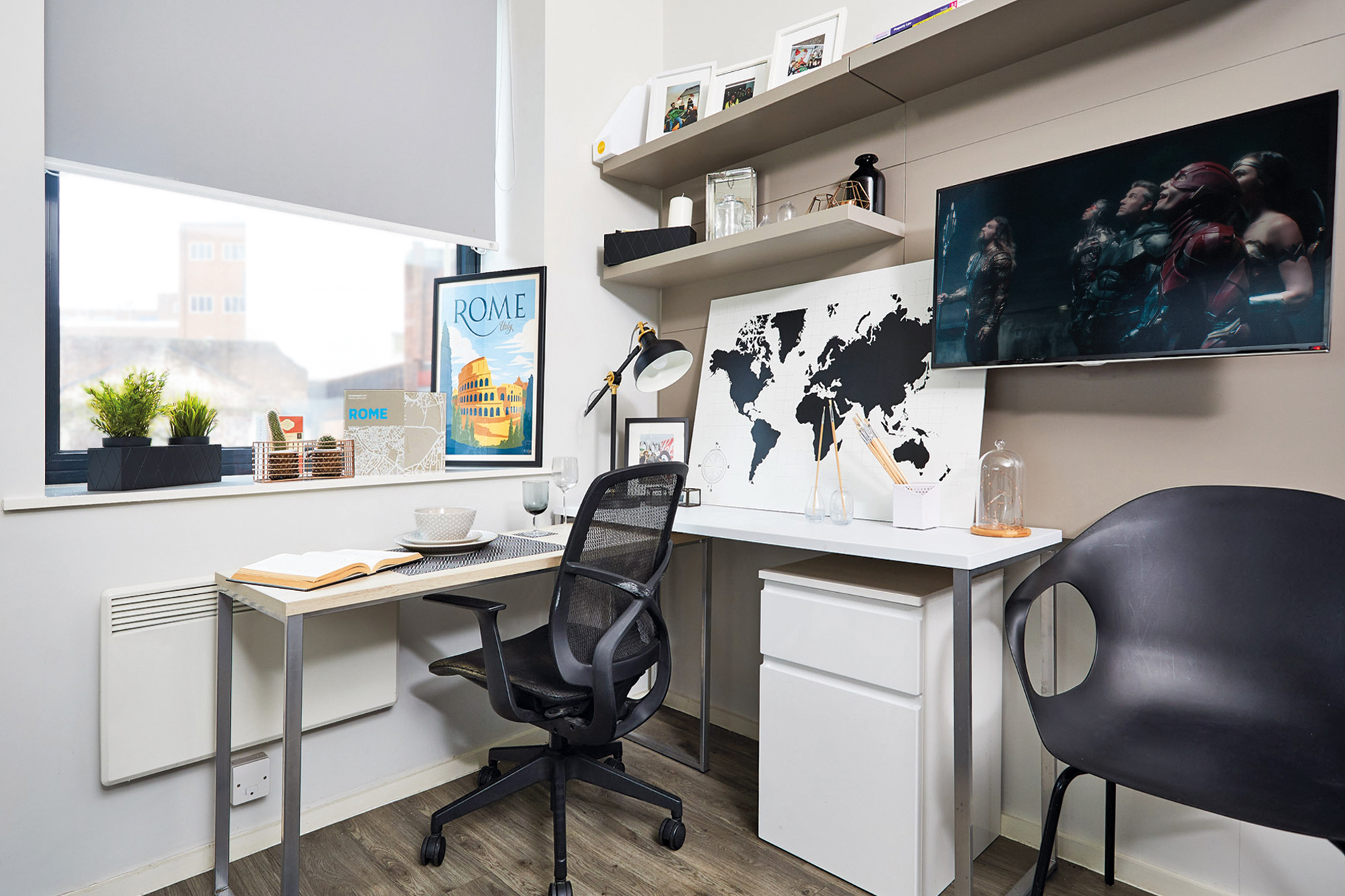 A modern home office in a Portland House features a desk with a black chair. A map and TV adorn the wall, alongside decorative shelves and a plant by the window. On the desk sit a book, plate, and teacup. A Rome poster leans stylishly as light floods through the large window.