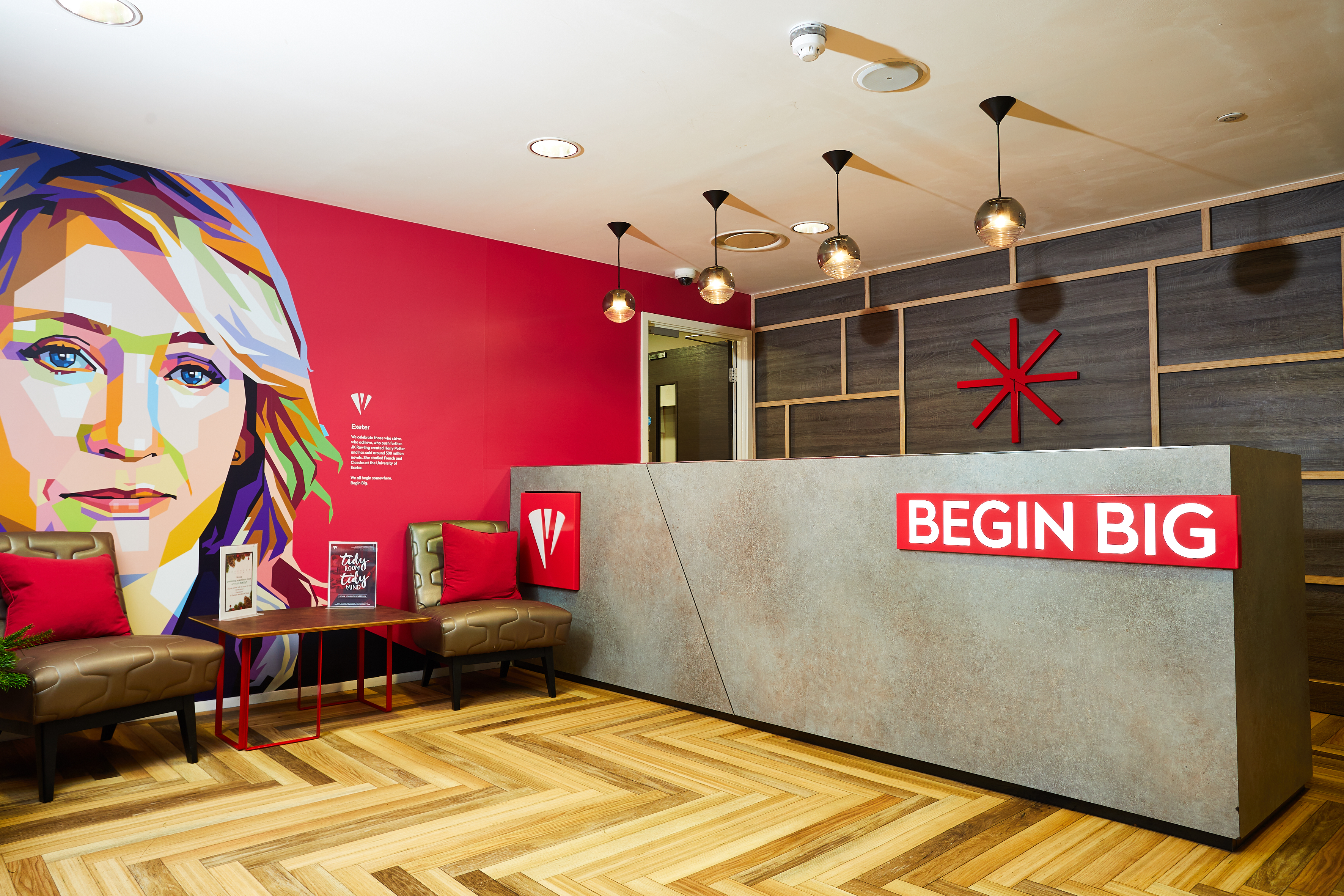The modern lobby of Portland House boasts a colorful wall portrait of a woman. The reception area features a large concrete desk with "BEGIN BIG" in bold red letters. Wooden flooring, green chairs, and hanging lights all come together to create a vibrant atmosphere.