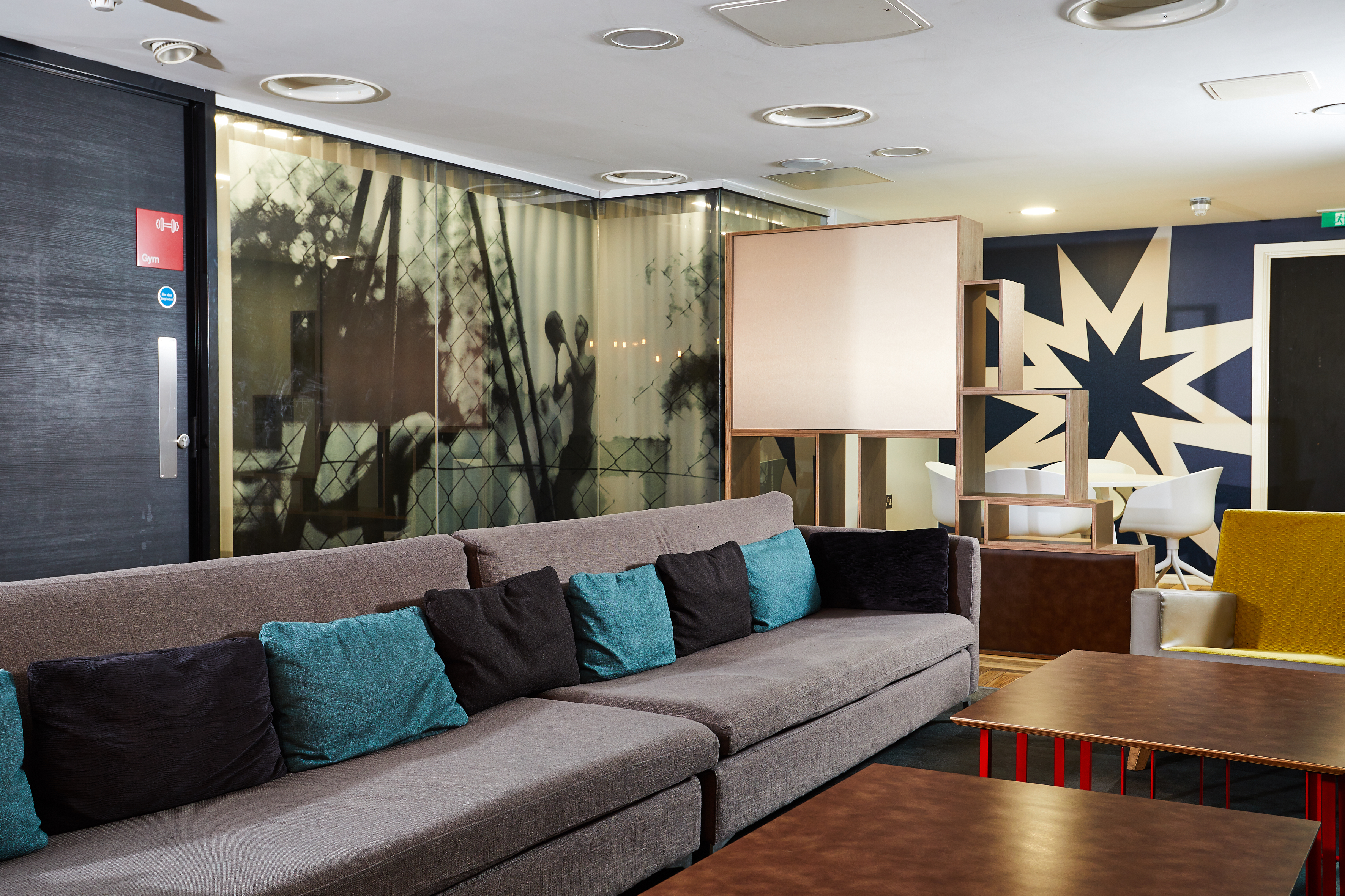 The Portland House's modern lounge area features a long gray sofa adorned with teal and black cushions. Wooden tables sit in front, while chairs rest against a backdrop of a large star design on the wall. Soft lighting and abstract art enhance the decor.