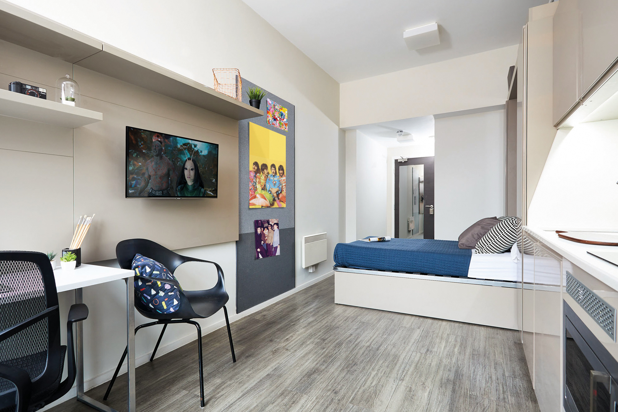 This Portland House studio boasts a modern design with a cozy bed, wall-mounted TV, and compact kitchenette. A study area with a desk and chair sits to the left. The decor features vibrant wall art and shelves, complemented by wood-style flooring and neutral-toned walls.