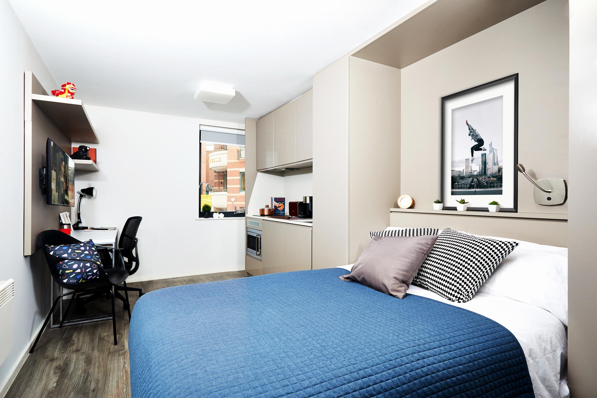 This modern Portland House bedroom features a blue bedspread, gray and checkered pillows, and built-in cabinetry. A desk with a chair and computer screen sits by the window. Wall art and small plants add charm to the space, which is completed with elegant wood flooring.