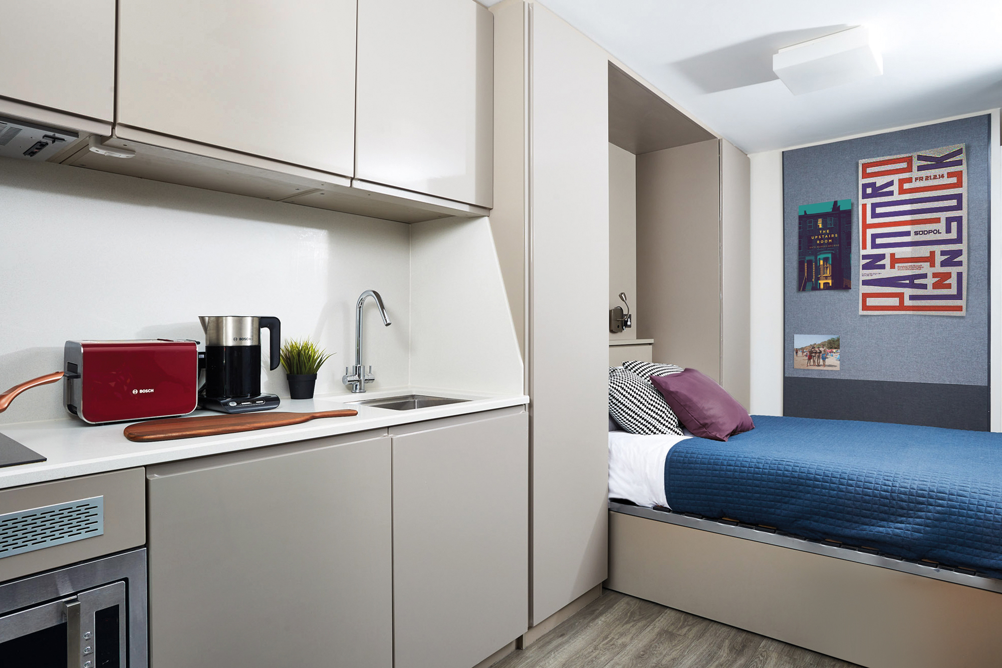 Modern studio apartment in Portland House with a compact kitchen featuring a red toaster, kettle, and sink. Adjacent is a sleeping area with a bed, blue bedding, and decorative pillows. Artwork and a small plant add a touch of color and life to the space.