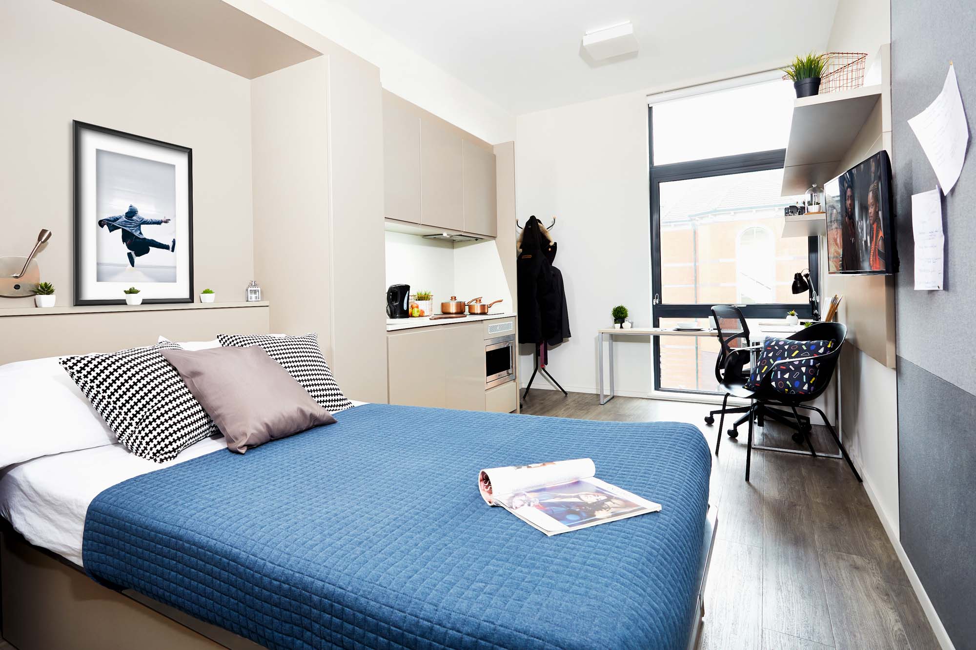 Modern studio apartment in Portland House with a cozy bed featuring a blue quilt, decorative pillows, a small kitchenette, and a workspace with a chair and desk. A coat hangs on a rack, and a large window lets in natural light. Art decorates the walls.