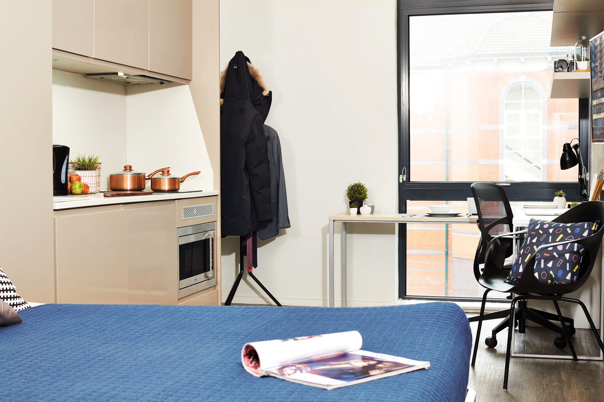Cozy studio apartment with a bed covered in a blue quilt, a small kitchen area featuring copper cookware, and a desk with a chair. The window offers a view of the brick Portland House building. A coat hangs on a stand near the kitchenette, adding an inviting touch.