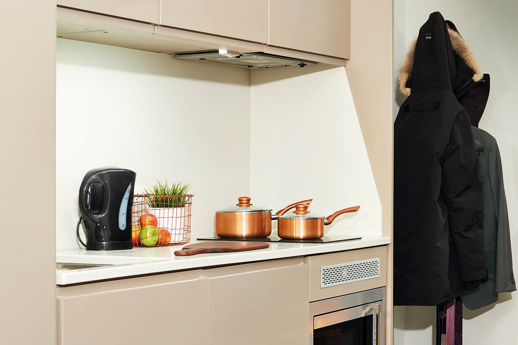 In a charming Portland house, the modern kitchen showcases a countertop adorned with a black electric kettle, a small plant, apples, and a cutting board. Copper pots rest on the stove, while coats hang neatly to the right. Neutral-toned cabinetry and a stainless steel oven complete the scene.