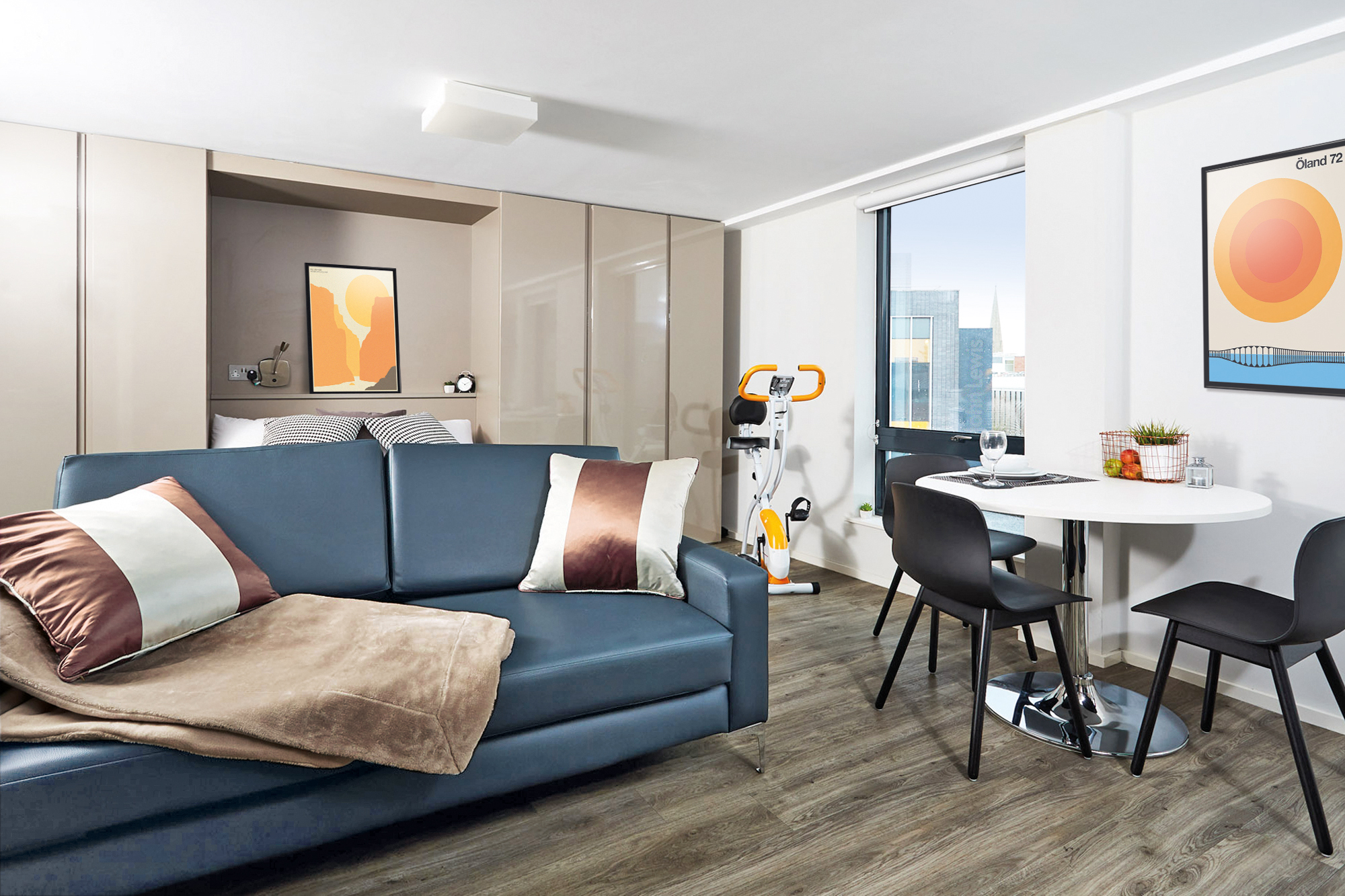 Modern studio apartment in Portland House with a blue sofa and cushions, a blanket, a round dining table with two chairs, an exercise bike, and artwork on the wall. A floor-to-ceiling window offers a cityscape view. Bedroom area partially visible.