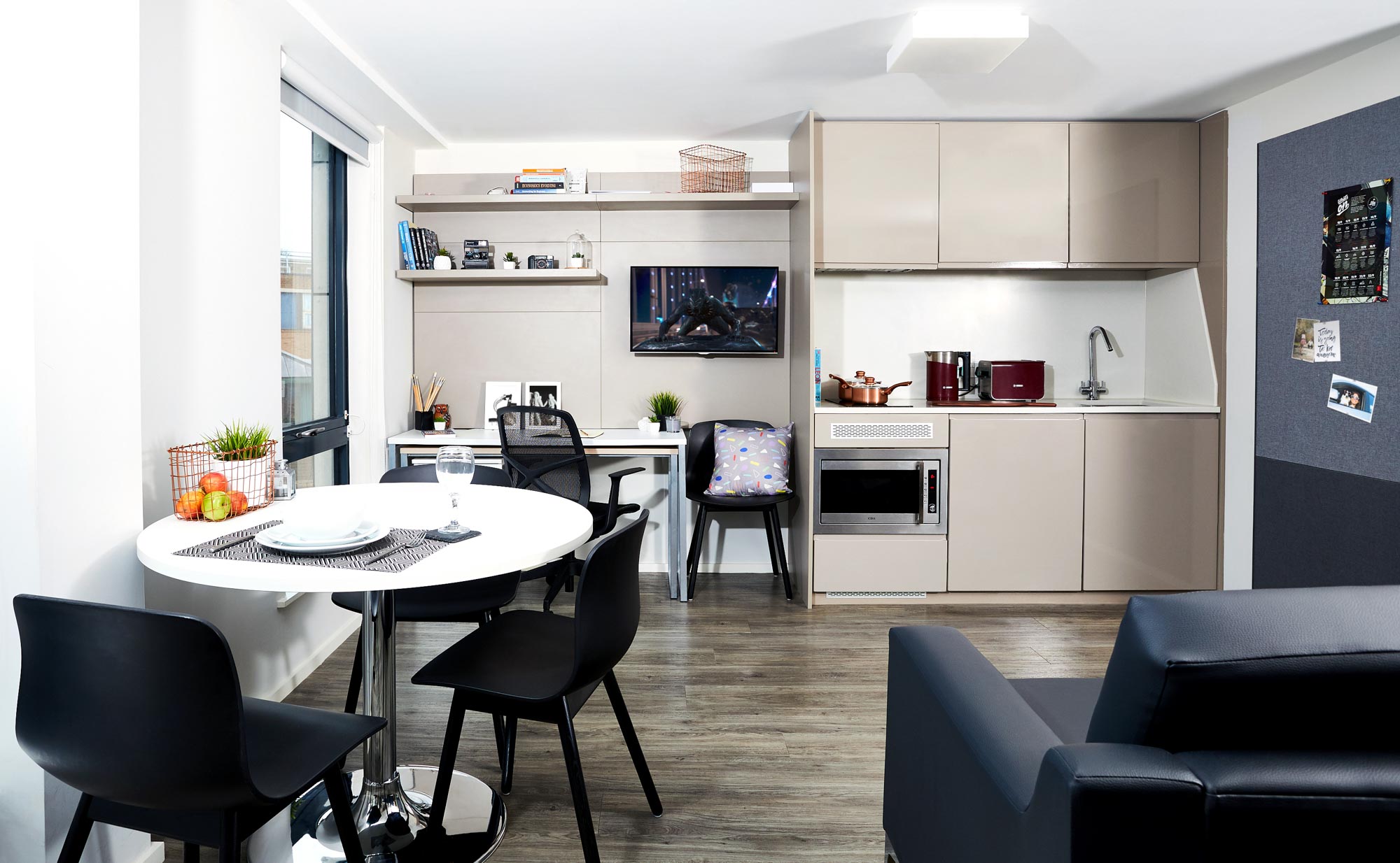 Modern studio apartment in Portland House with a small kitchen, dining table for two, and a cozy workspace. The kitchen features light cabinets, a sink, and appliances. A sofa sits on the wooden floor, and shelves hold decorative items that add charm to this inviting space.