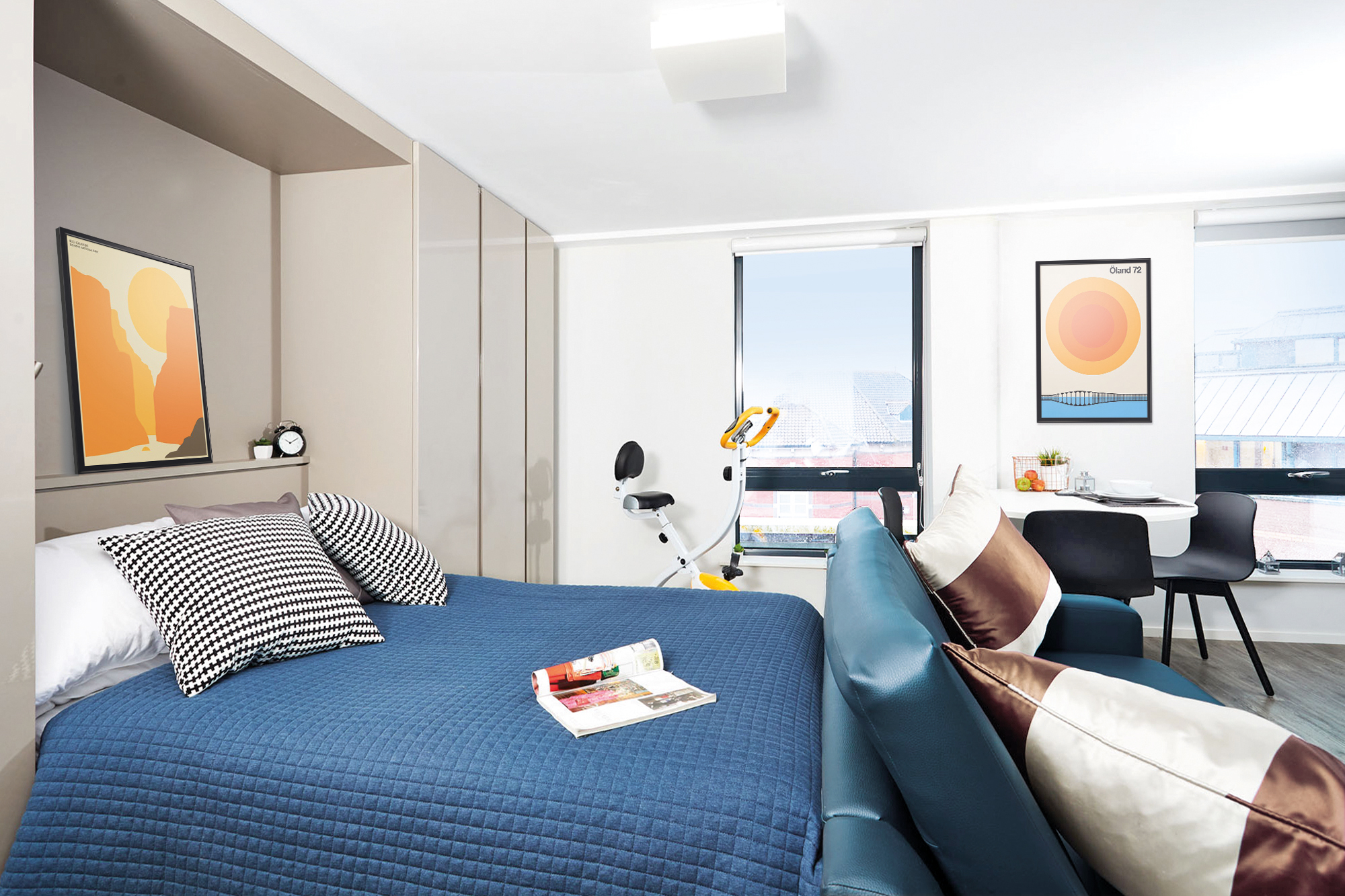 In this Portland House bedroom, a modern aesthetic prevails with a blue quilted bed and striped pillows. A bicycle and desk with chairs sit by the window. Abstract art graces the white walls, while a magazine on the bed and clock on the bedside shelf add personal touches.