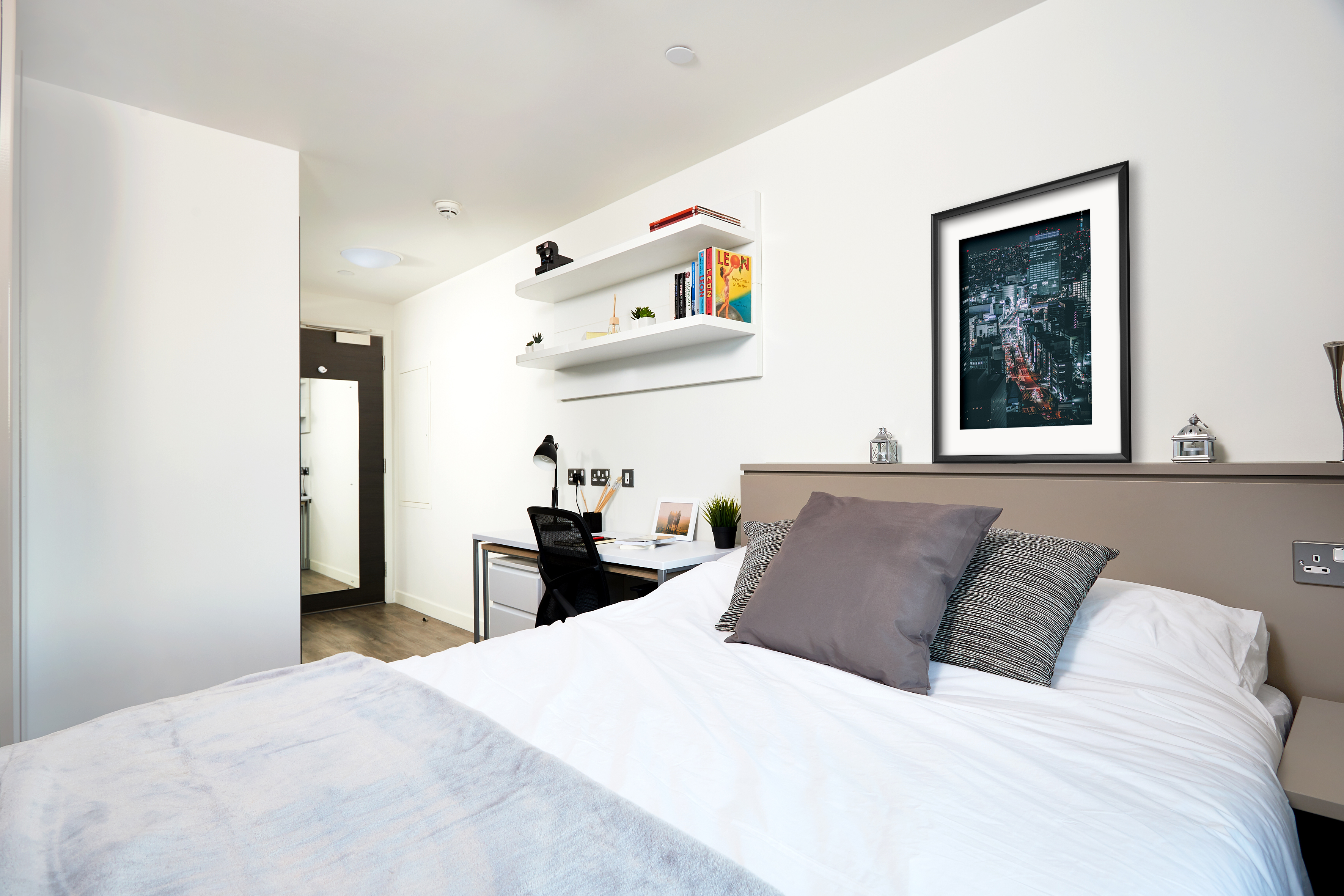 A tidy bedroom features a neatly made bed with gray and white pillows, a framed cityscape on the headboard, and white shelves above a desk. The room has white walls and an open door leading to a hallway.