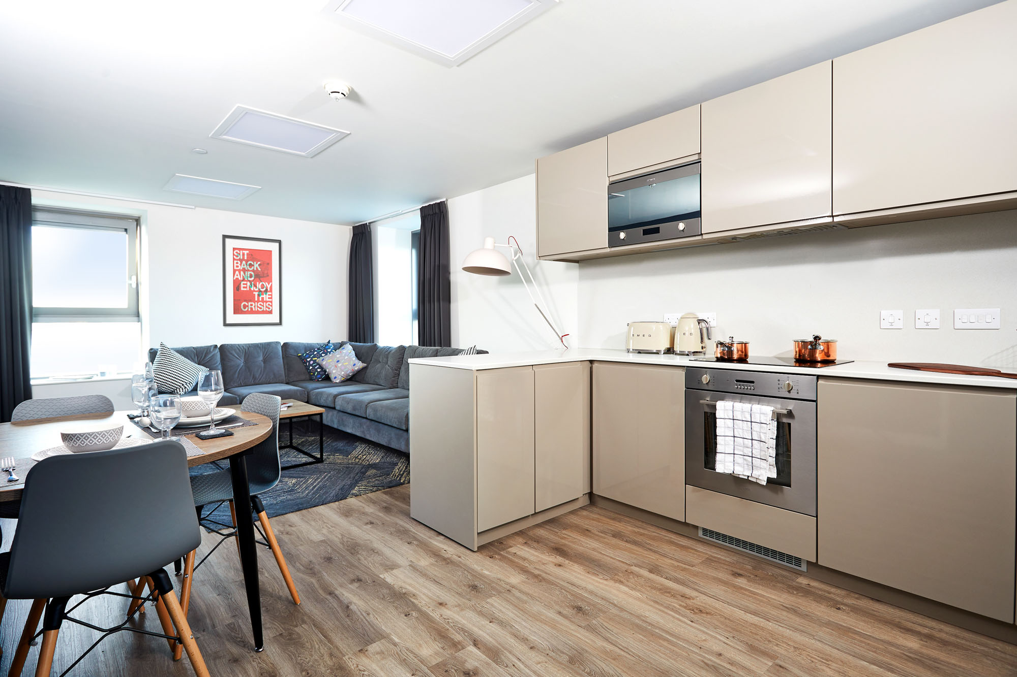 Modern kitchen and living area with light wood floors. The kitchen features beige cabinets, an oven, and a microwave. A dining table with blue chairs sits nearby. The living area has a gray sofa and a wall poster. Large windows bring in natural light.