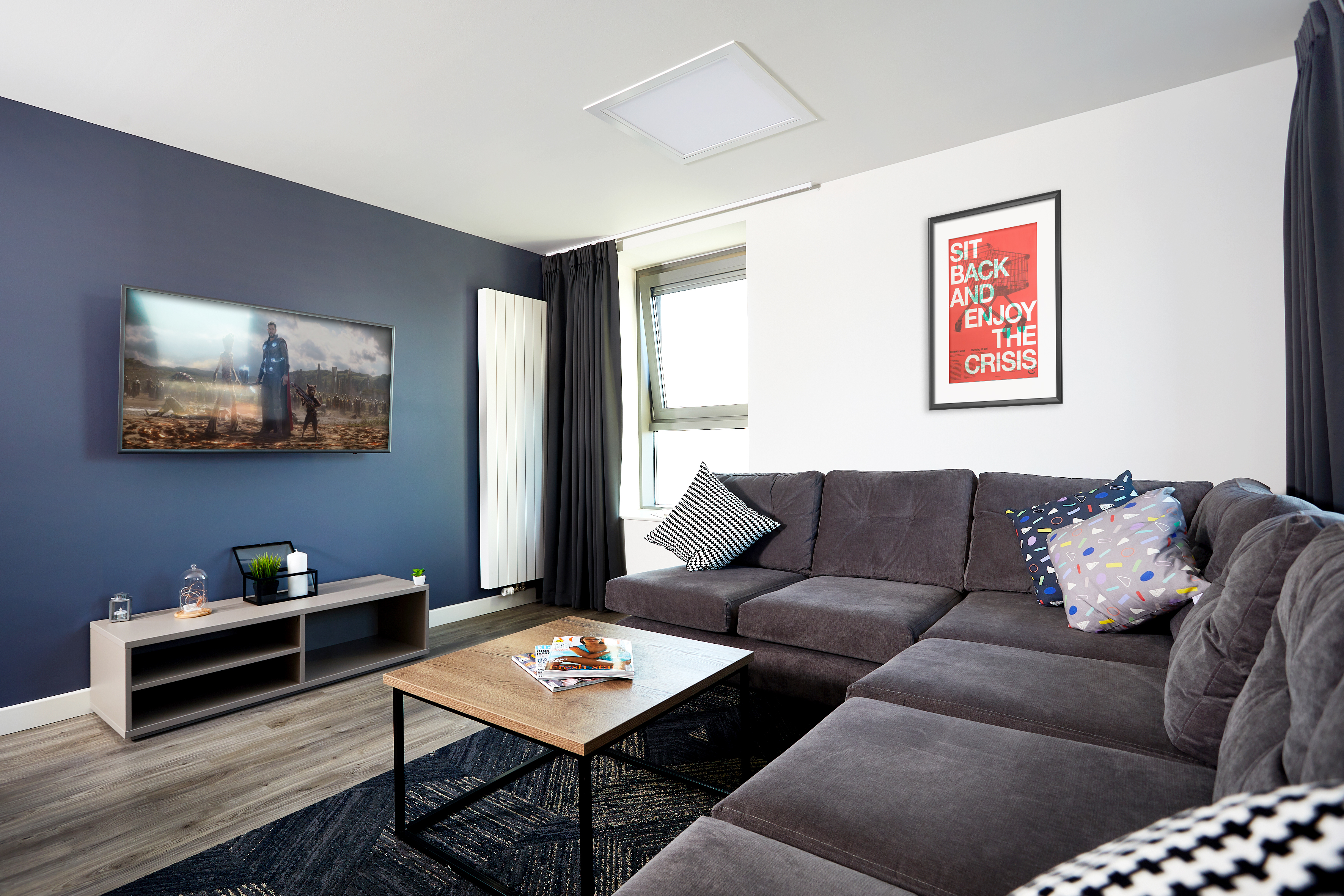 A modern living room with a gray sectional sofa, colorful pillows, and a wooden coffee table. A TV is mounted on a blue accent wall, and a red and black framed poster hangs nearby. A window with curtains lets in natural light.