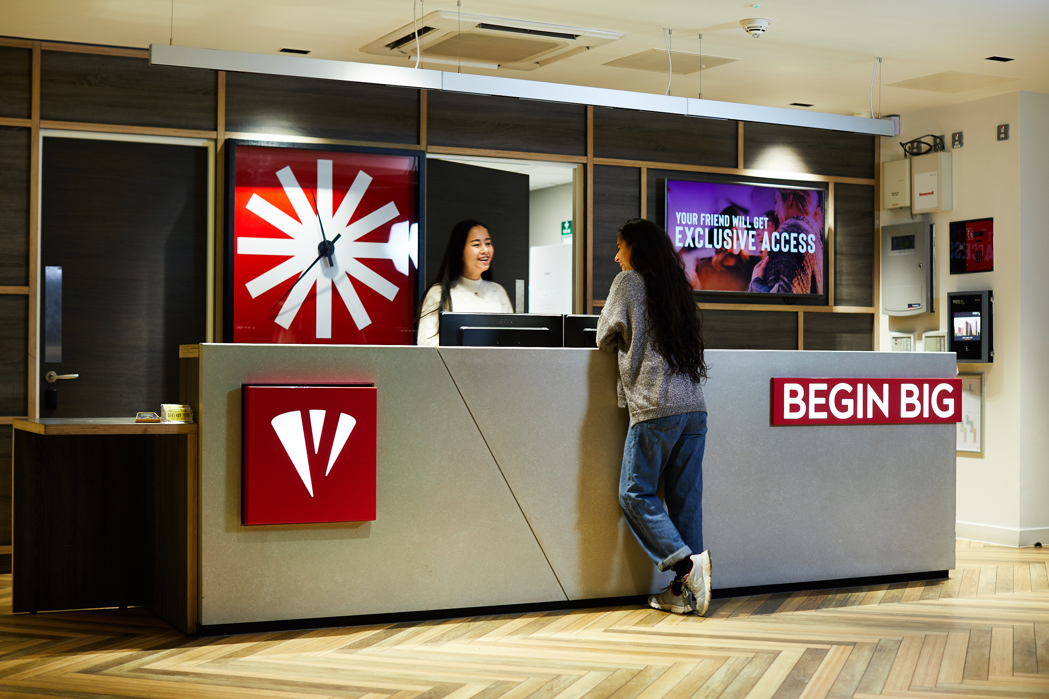 A person with long hair leans on a modern reception desk while speaking to another person behind it. The desk has the text "BEGIN BIG" and a red and white logo. A digital sign reads "YOUR FRIEND WILL GET EXCLUSIVE ACCESS." The floor is herringbone wood.