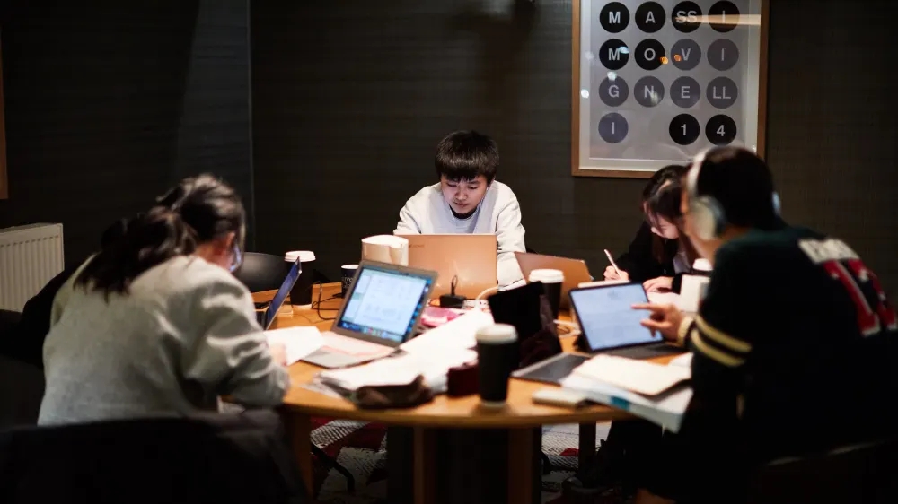 A group of people working at a round table, each focused on their laptops and notes, embodying effective study tips in the dimly lit room. A framed eye chart is visible on the wall in the background, guiding their focus as they emulate studying at home.