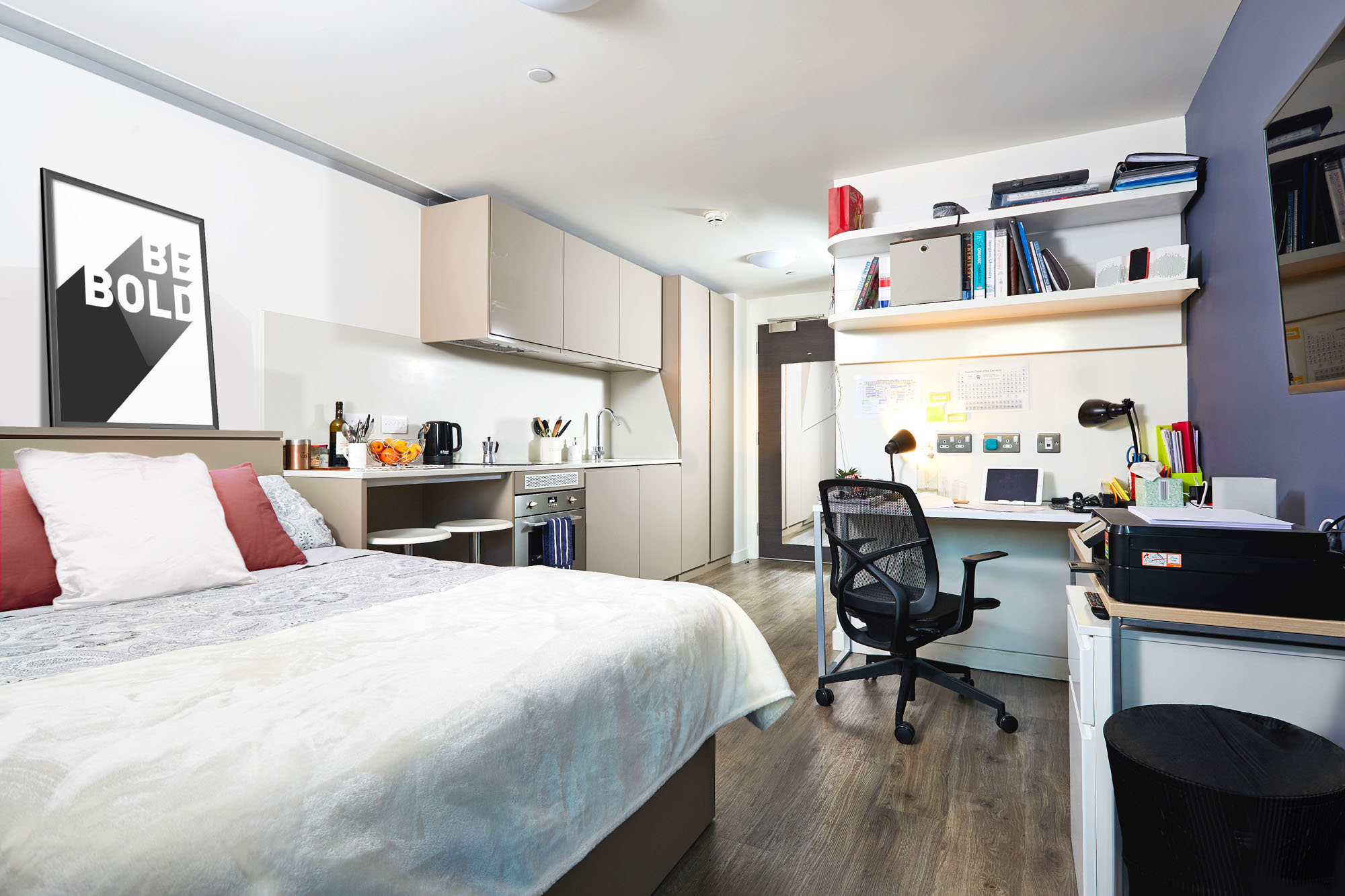 Modern studio apartment with wood flooring, featuring a neatly made bed, compact kitchen with appliances, and a desk with a chair and computer. Wall shelves hold books and decor, and a "Be Bold" poster adds a motivational touch. Bright and organized space.