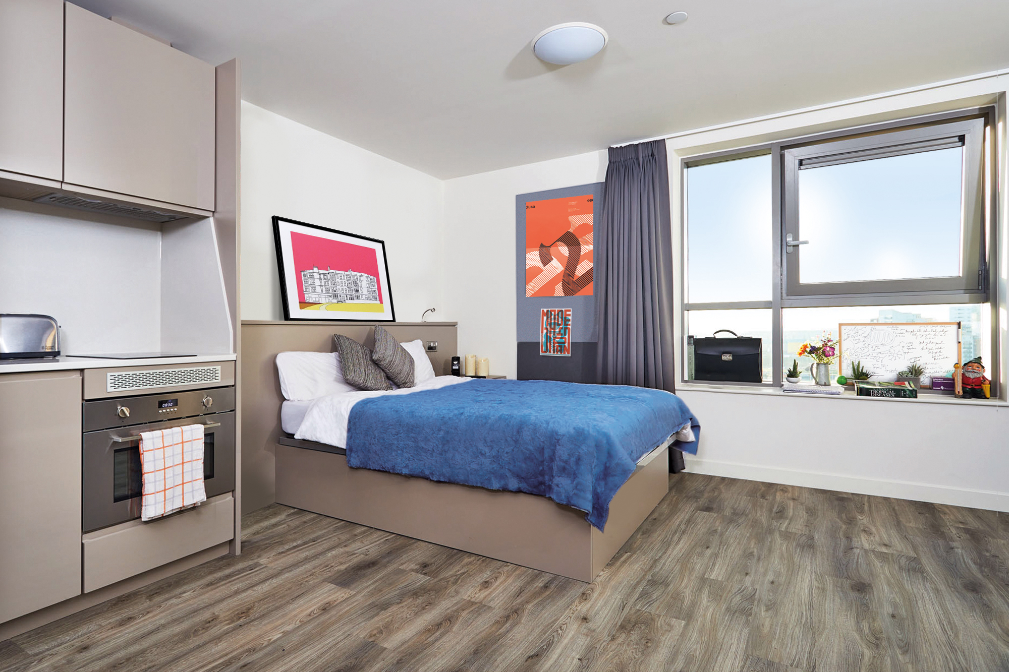 A modern studio apartment with a bed featuring a blue comforter, a small kitchen area with an oven and cabinets, and large window with a city view. Artwork decorates the walls, and the floor is wooden.