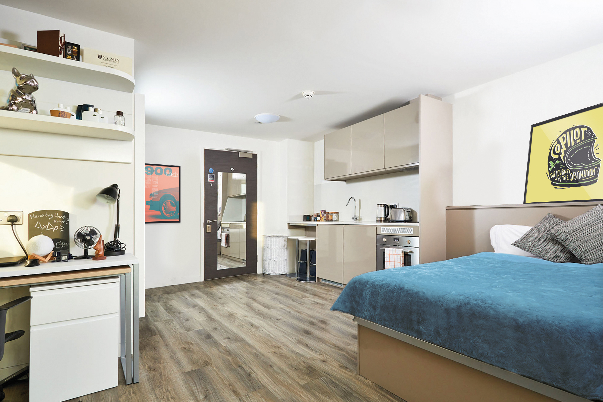 A modern studio apartment features a neatly made bed with blue bedding. The room includes a desk and shelves on one side, a compact kitchen, and bright wooden flooring. A large yellow poster adorns the wall above the bed.