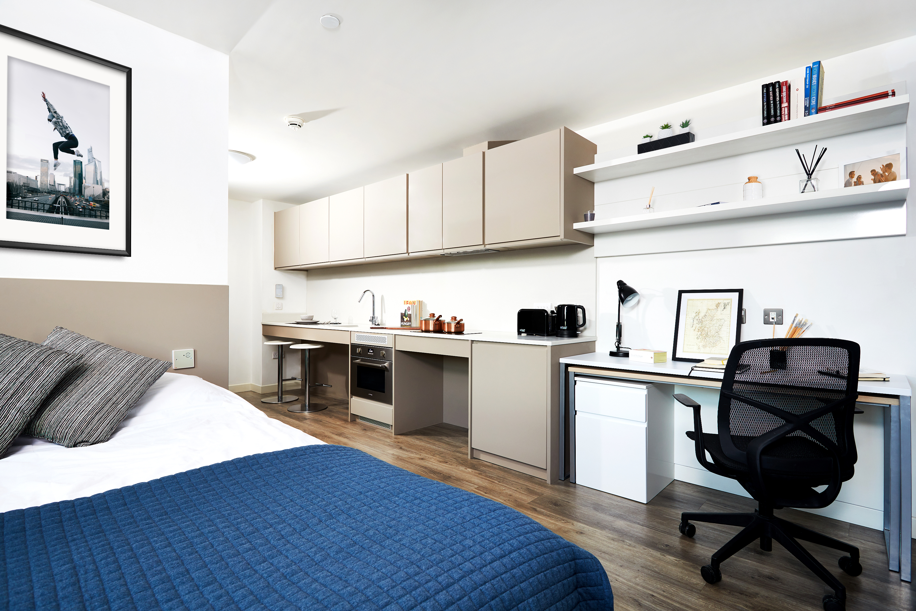 Modern studio apartment with a cozy bed, a small kitchenette with tan cabinets, and a desk area. The workspace includes shelves with books, a framed picture, a lamp, and a chair. The room is well-lit with wood flooring and a blue bed cover.