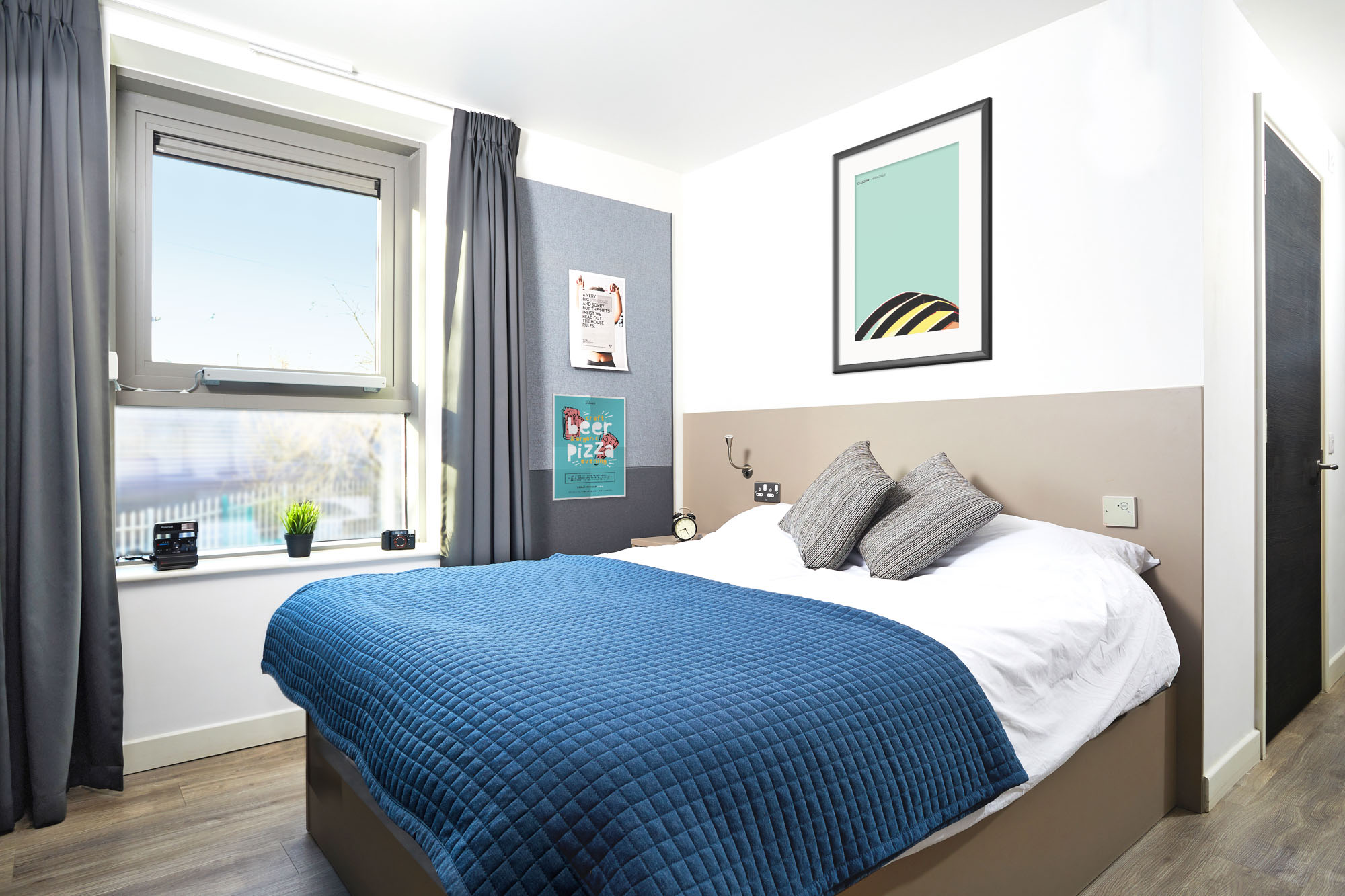 Modern bedroom with a double bed, blue quilt, and two gray pillows. A window with gray curtains is on the left. Wall art includes abstract prints and a pizza sign. A wooden floor and minimal decor create a clean, stylish look.