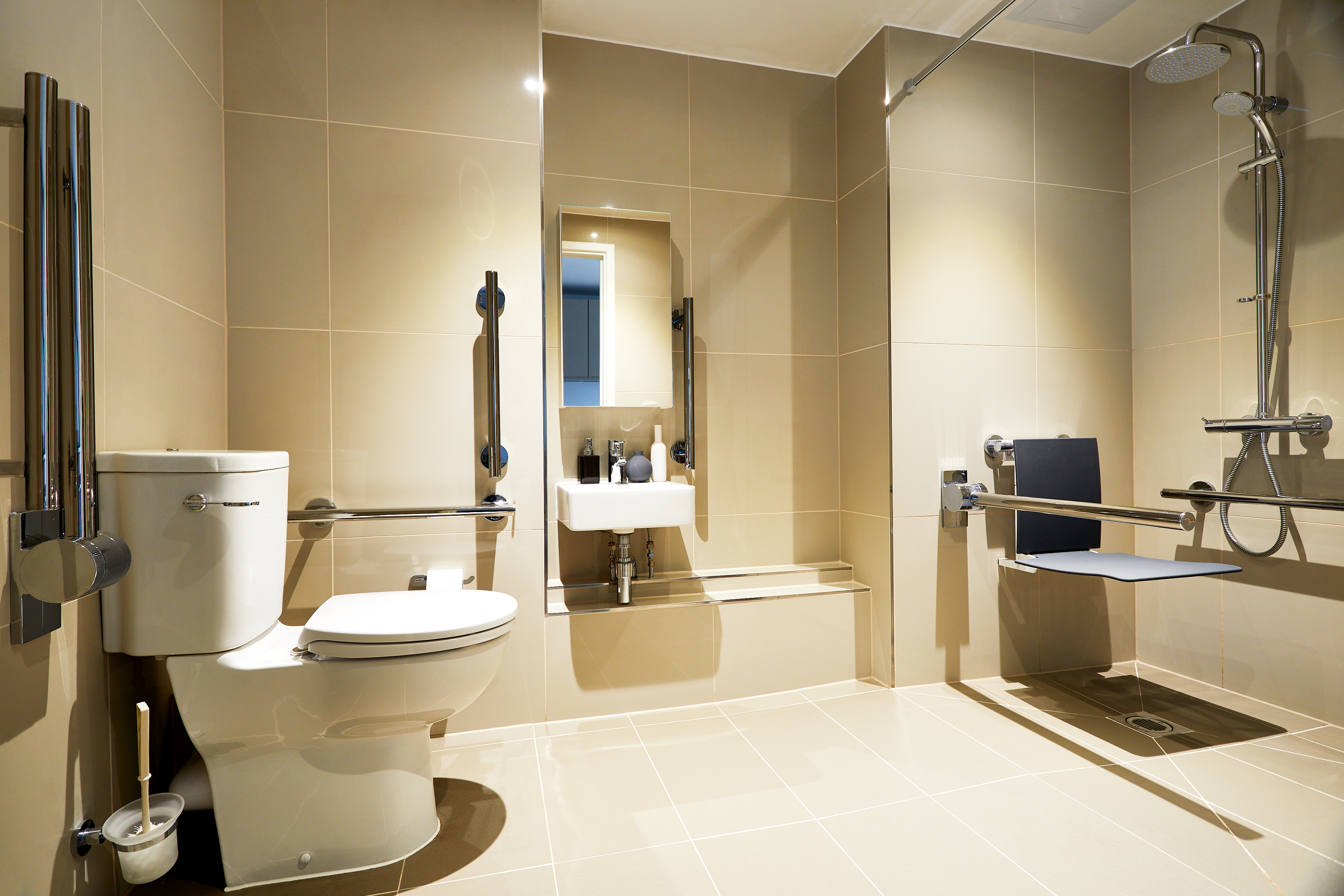 A modern, accessible bathroom with beige tiles features a toilet with support rails, a sink beneath a mirror, and a shower area with a folding seat and additional handrails for support.