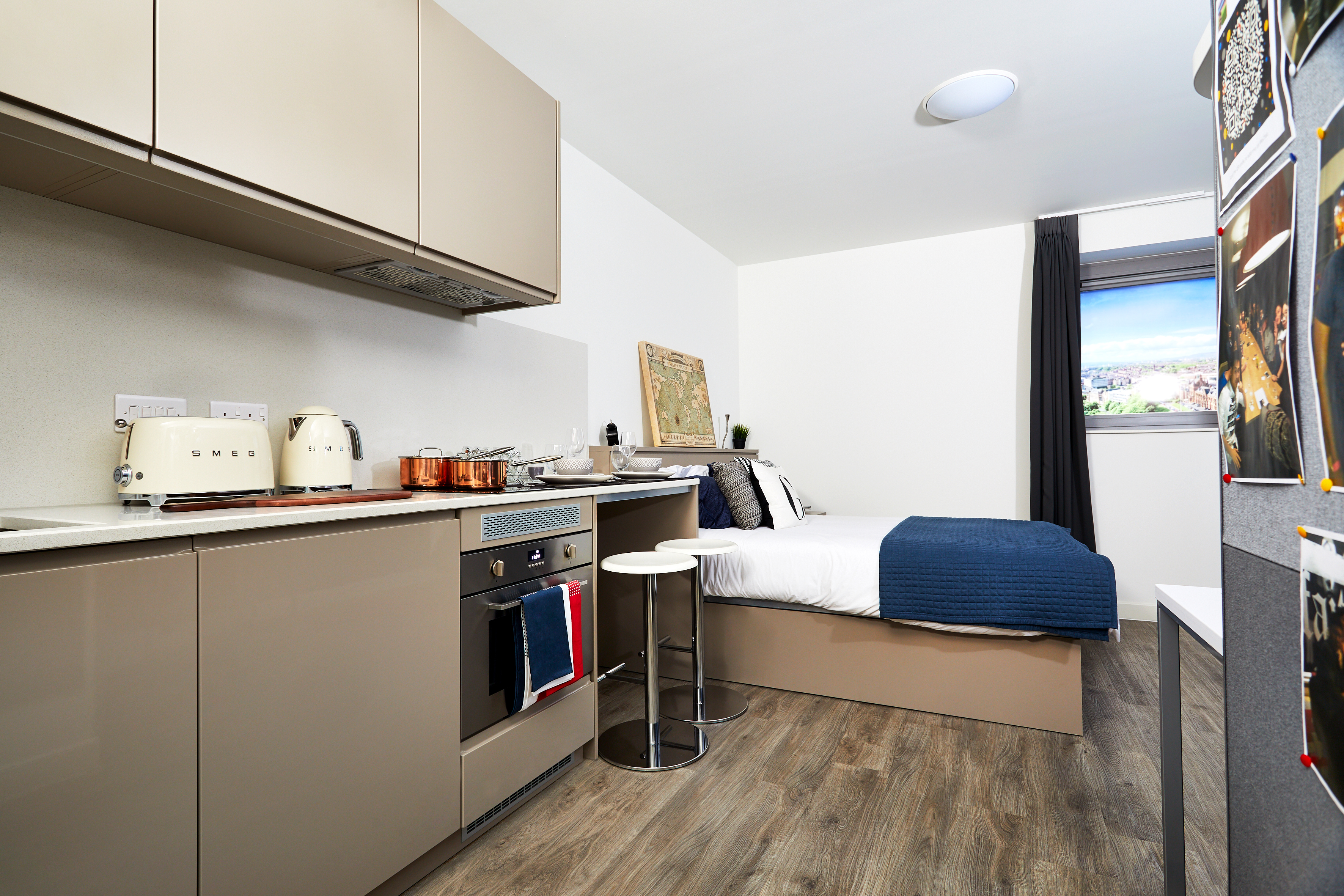 Modern studio apartment with a compact kitchen featuring beige cabinets, an oven, kettle, toaster, and cookware. Adjacent is a cozy bed with blue bedding, a side table, and a large window. Photographs decorate the wall, and the floor is wooden.
