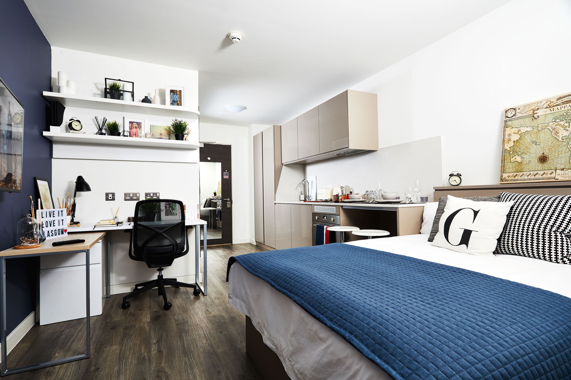 Modern studio apartment with a bed, blue blanket, and decorative pillows. A desk with a chair, lamp, and laptop is against a dark wall. Shelves with plants and decor are above. A small kitchen area and a world map on the wall complete the cozy setup.